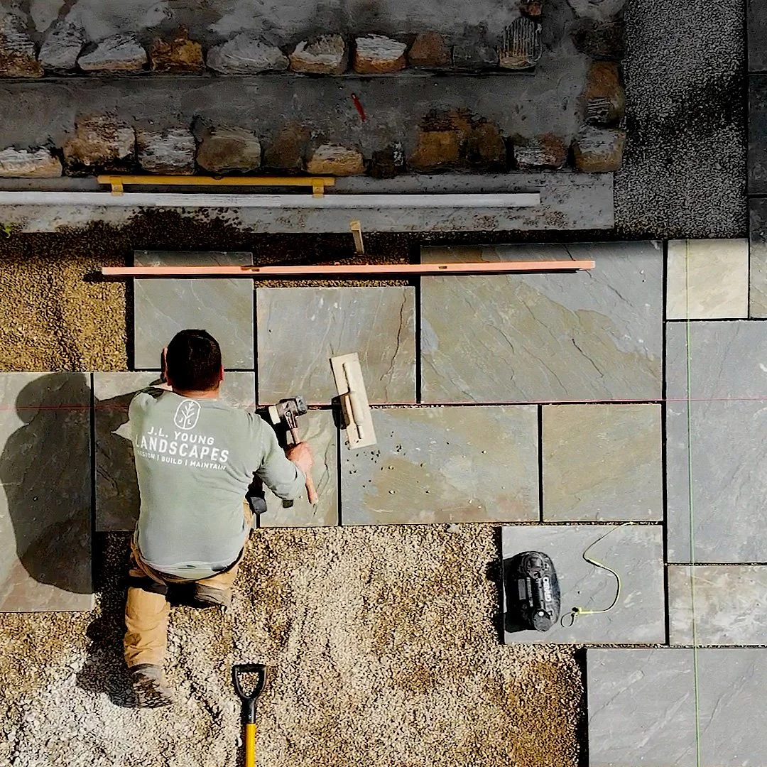 A worker installing large stone tiles on the ground, with some tiles already in place, using a power tool. The worker is kneeling on a gravel surface, wearing a shirt that reads, JL Young Landscapes'.