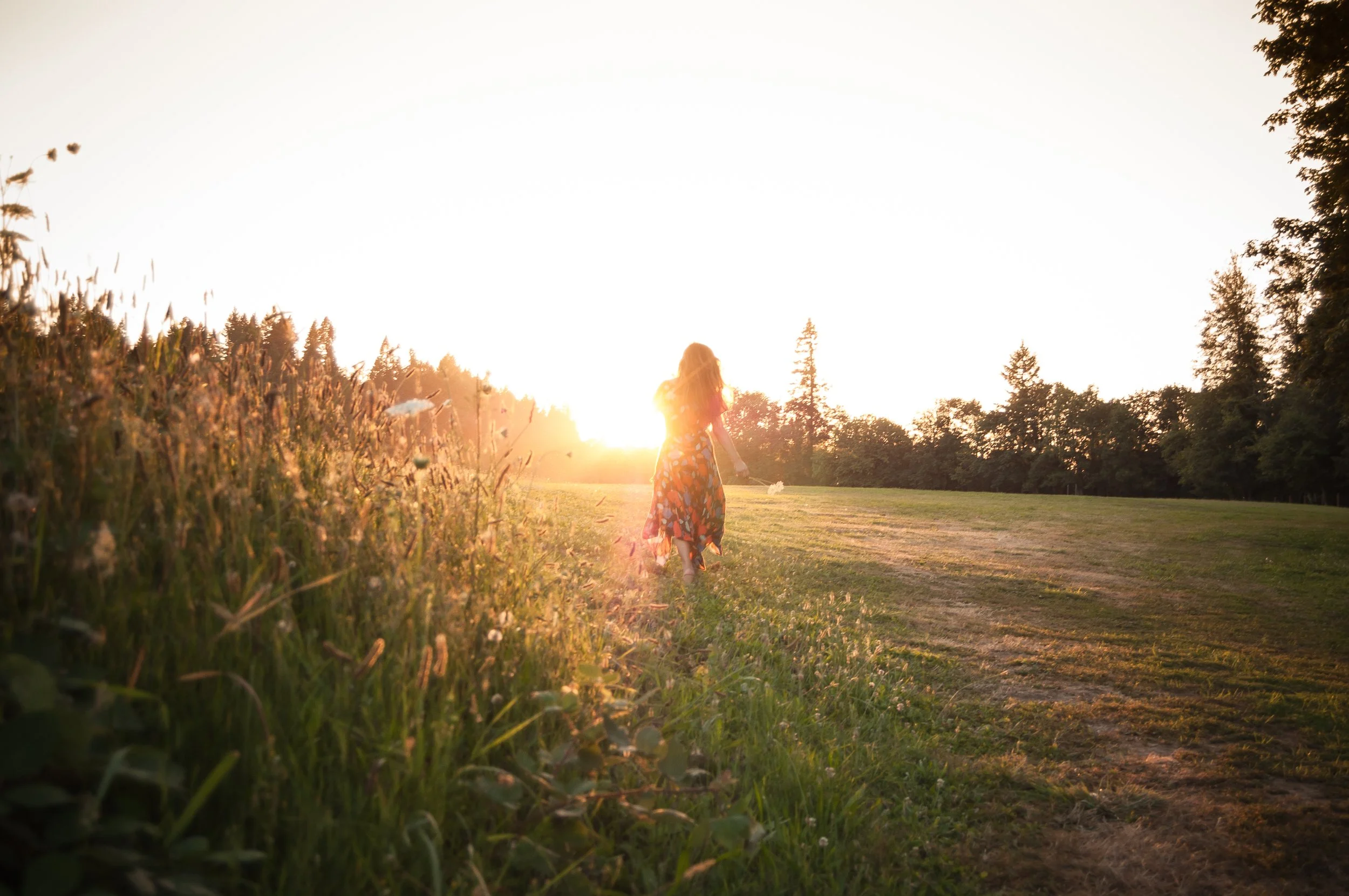 A girl walking through a grassy field at sunset, with trees in the background and sunlight creating a warm glow.