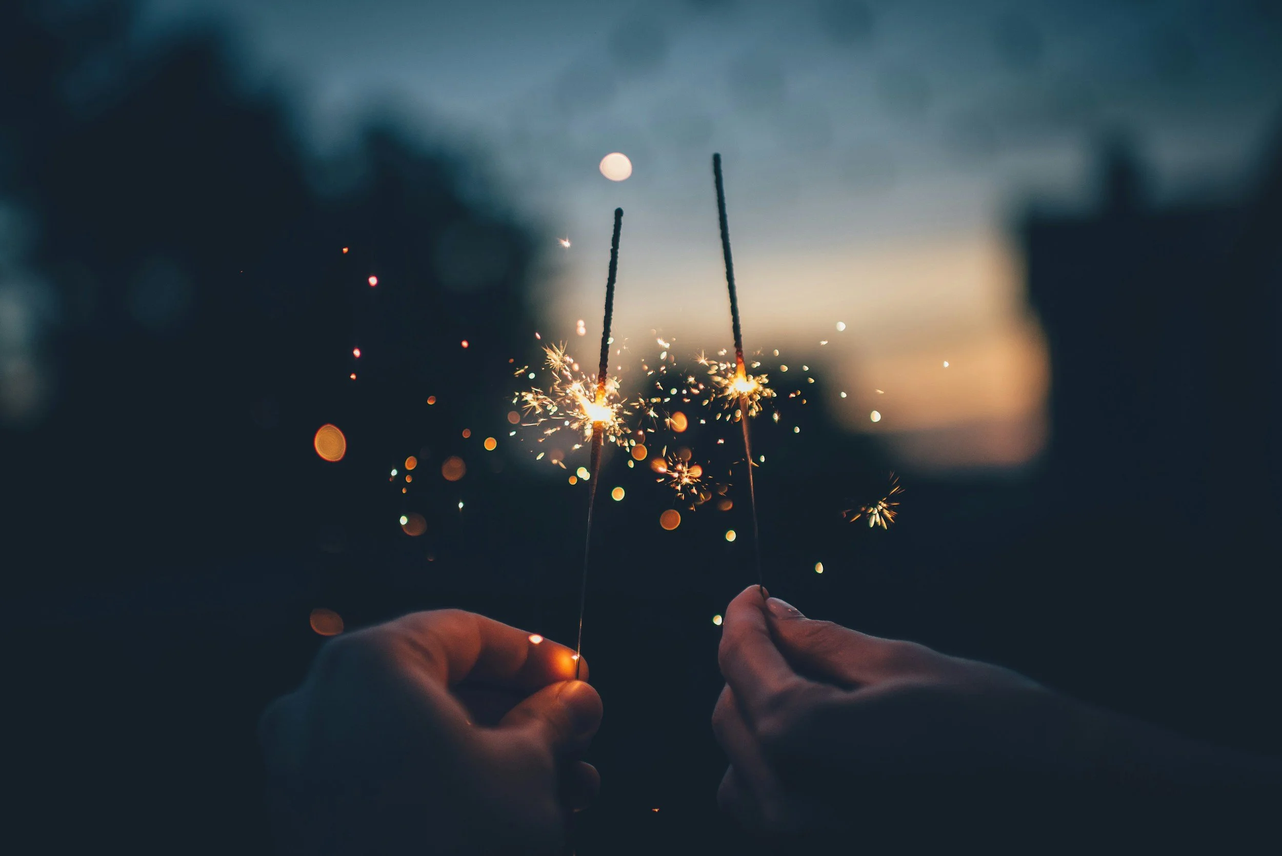 Two hands holding sparklers outdoors during dusk or night, with sparks flying and a blurred background featuring a full moon in the sky.