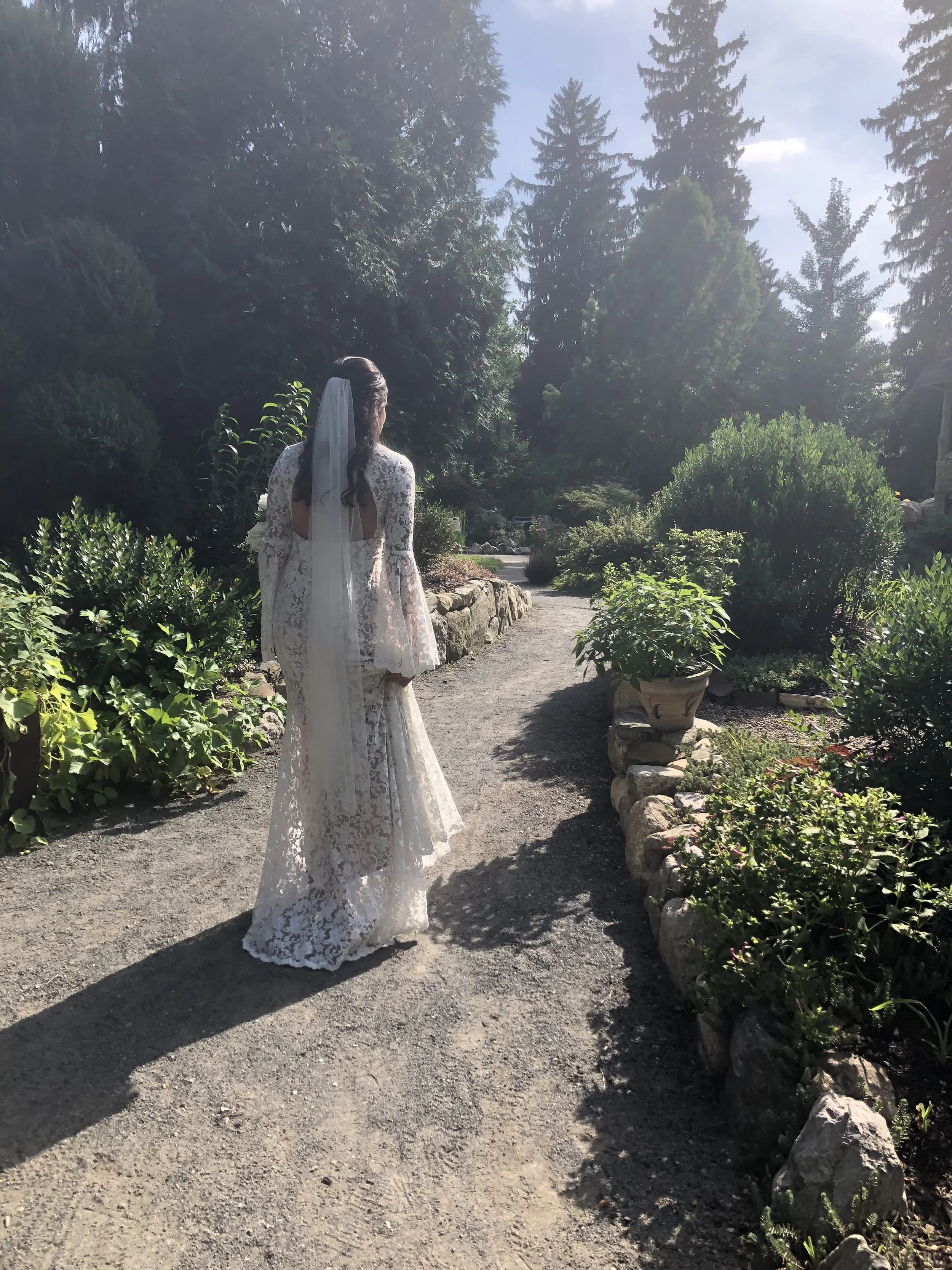A woman in a white lace wedding dress standing on a garden path surrounded by lush greenery and trees, with sunlight filtering through the leaves.