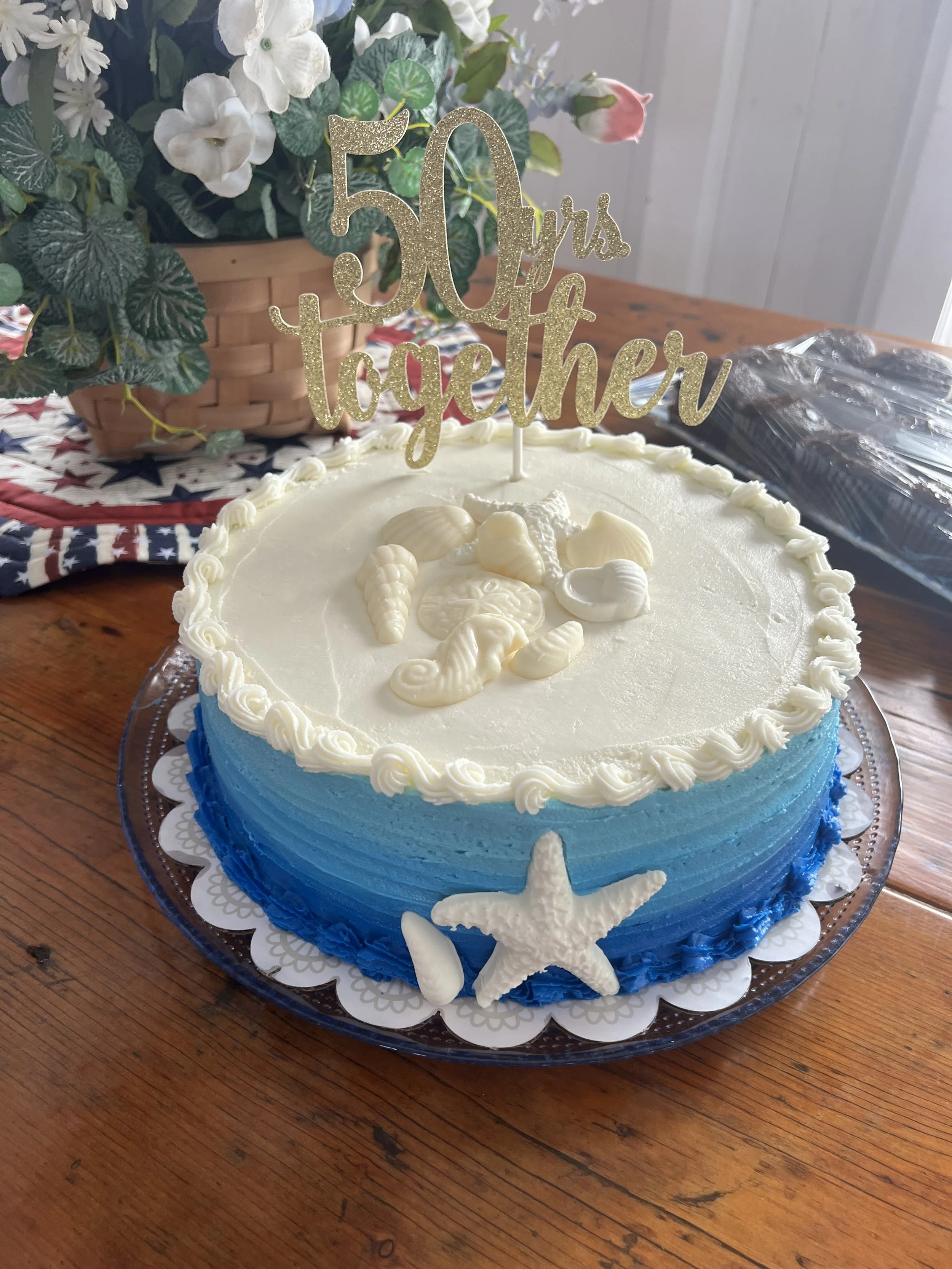 A round cake decorated with white frosting, shell and starfish edible decorations on top, and blue frosting around the sides. A gold glittery topper reads '50 yrs together.' The cake is on a silver tray on a wooden table, with a potted plant with white flowers in the background.