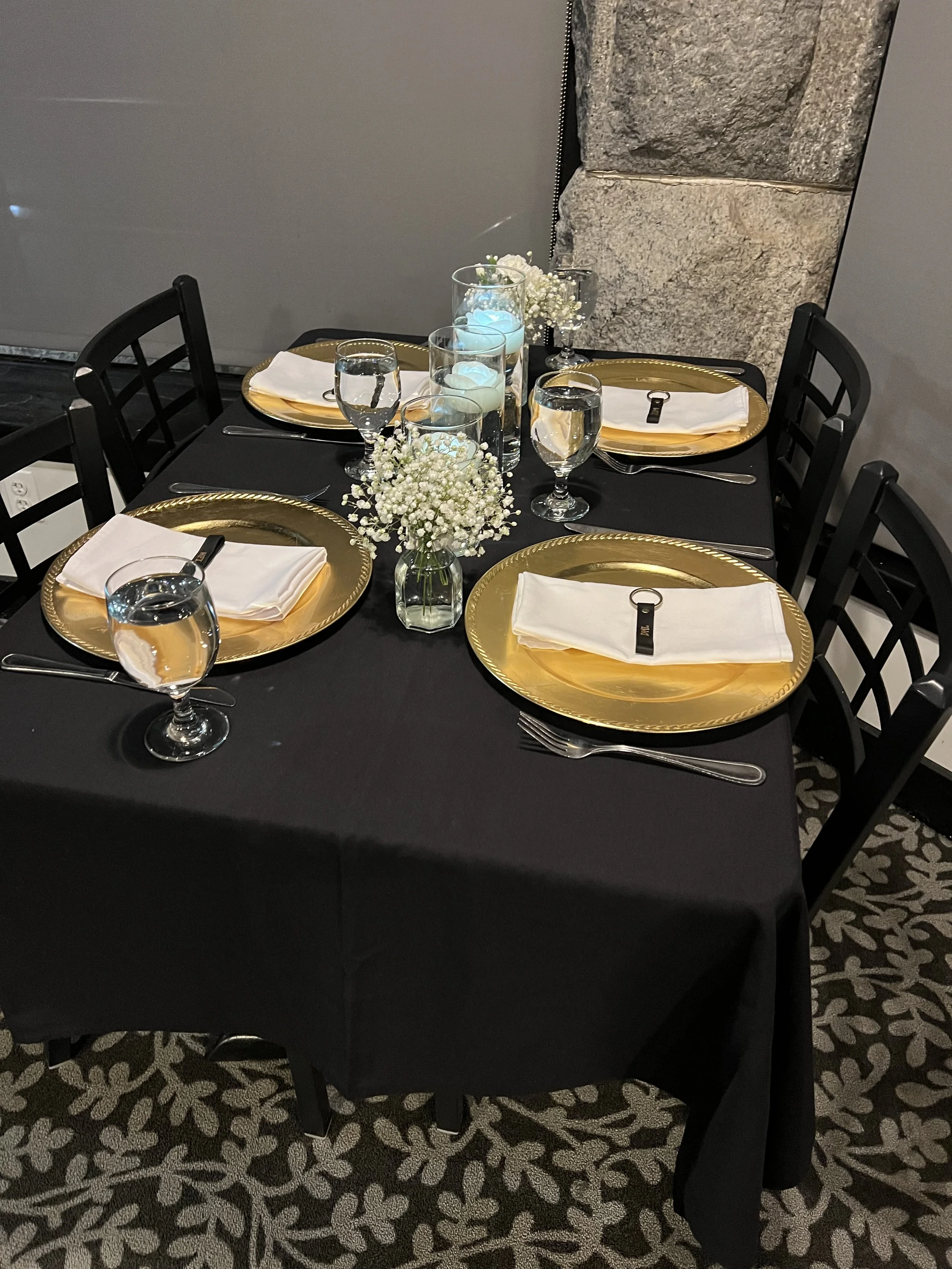 A dining table set for four with gold chargers, white napkins, and glasses of water. In the center are tall vases with white flowers and floating candles. The tablecloth is black, and the chairs are black with a lattice back design. The background features a marble wall and a patterned carpet.