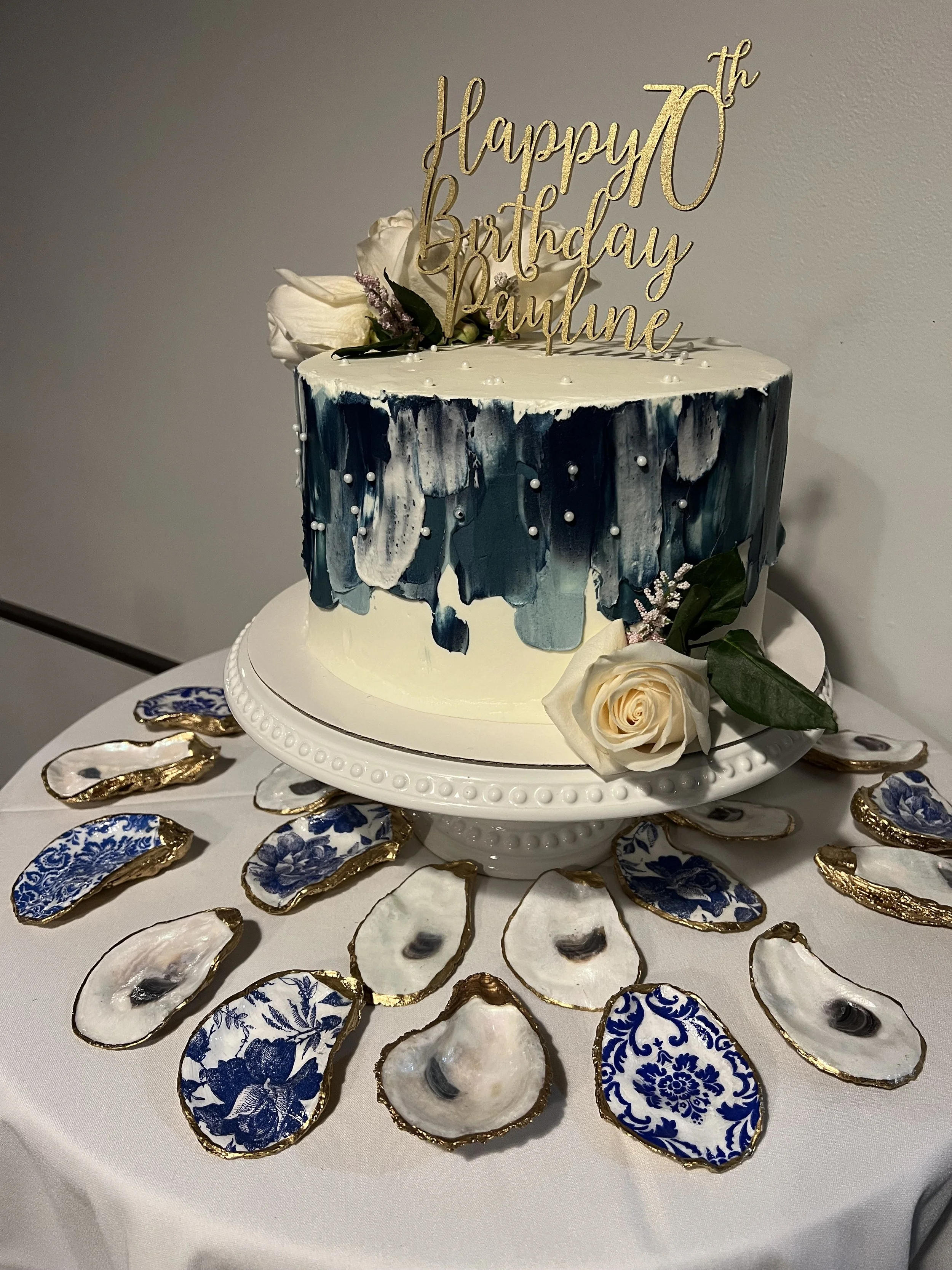 A decorated cake with a gold 'Happy 70th Birthday Pauline' topper, white roses, and purple accents. The cake is on a white cake stand surrounded by blue and white porcelain oyster shells with gold edges, arranged on a table with a white tablecloth.
