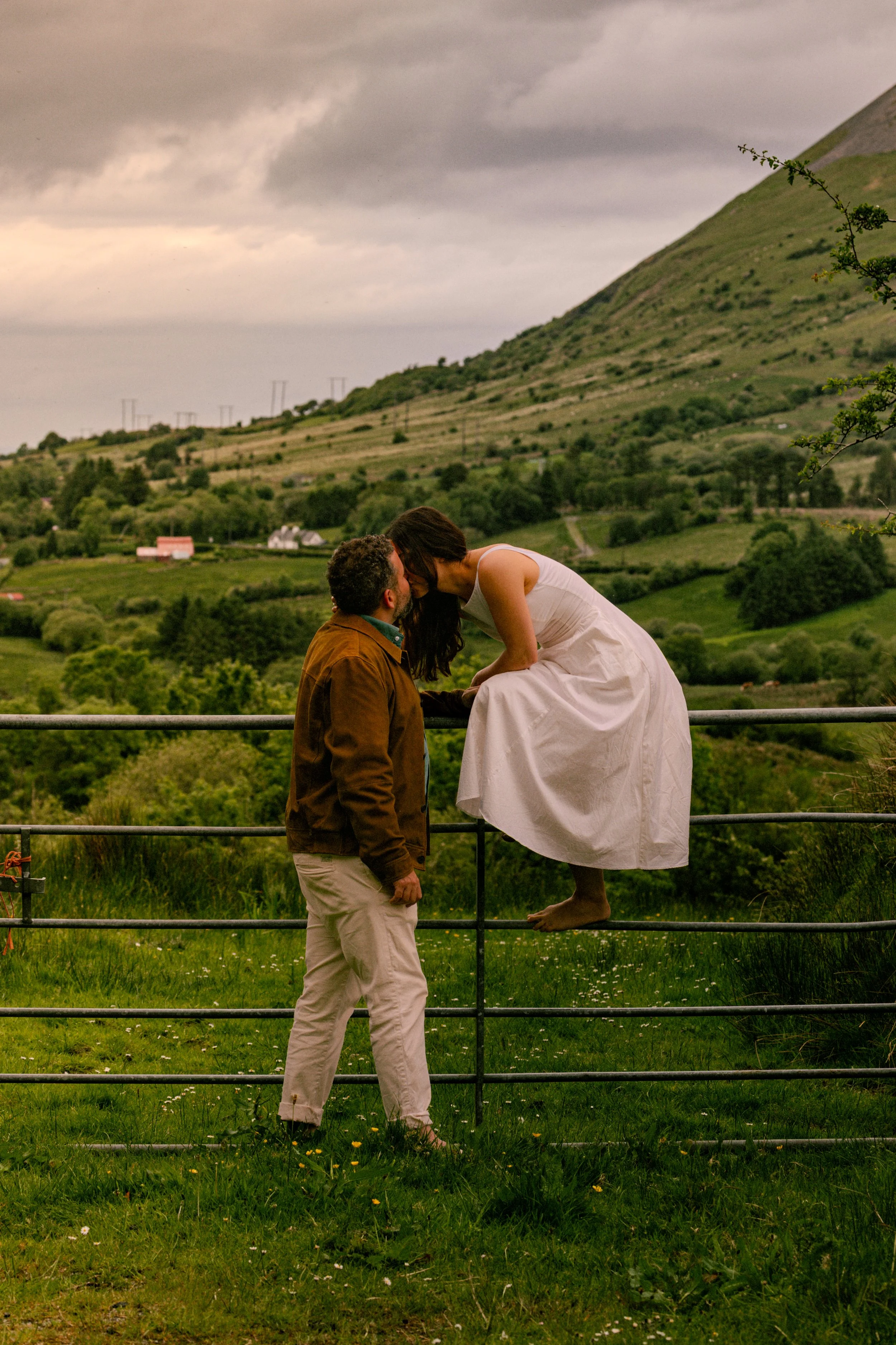A man and woman share a kiss outdoors, with the woman perched on a railing, against a lush green hillside with cloudy sky in the background.