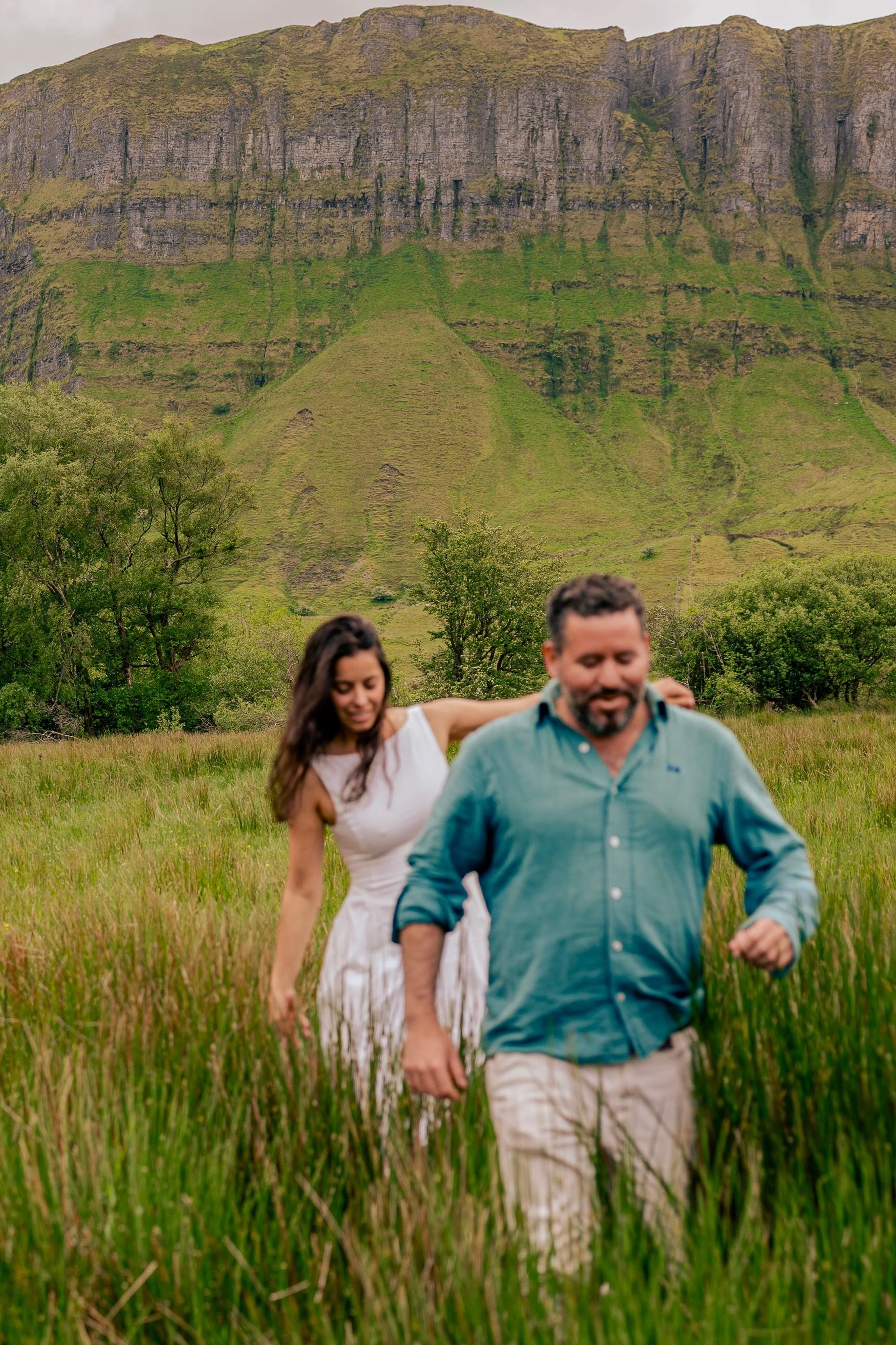 Two people, a woman and a man, walking through a grassy field with a large green mountain in the background.