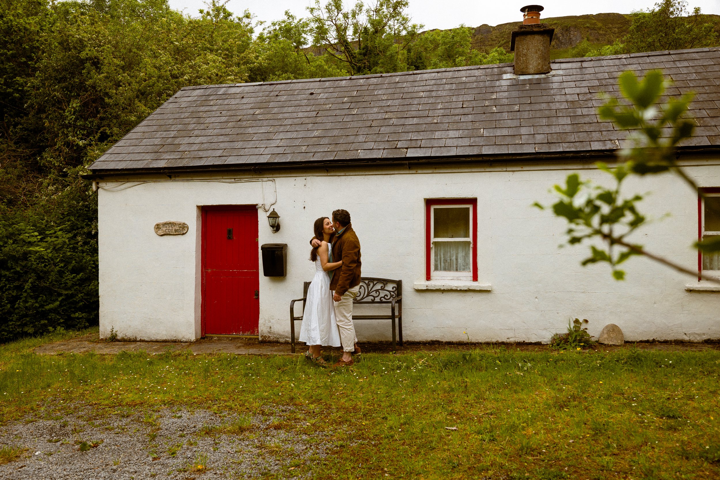 A couple stands in front of a small white house with a red door, hugging and smiling at each other. The house has a window with red trim, a bench, and a sign that reads 'Rose Cottage'. The roof is gray and surrounded by greenery and trees.
