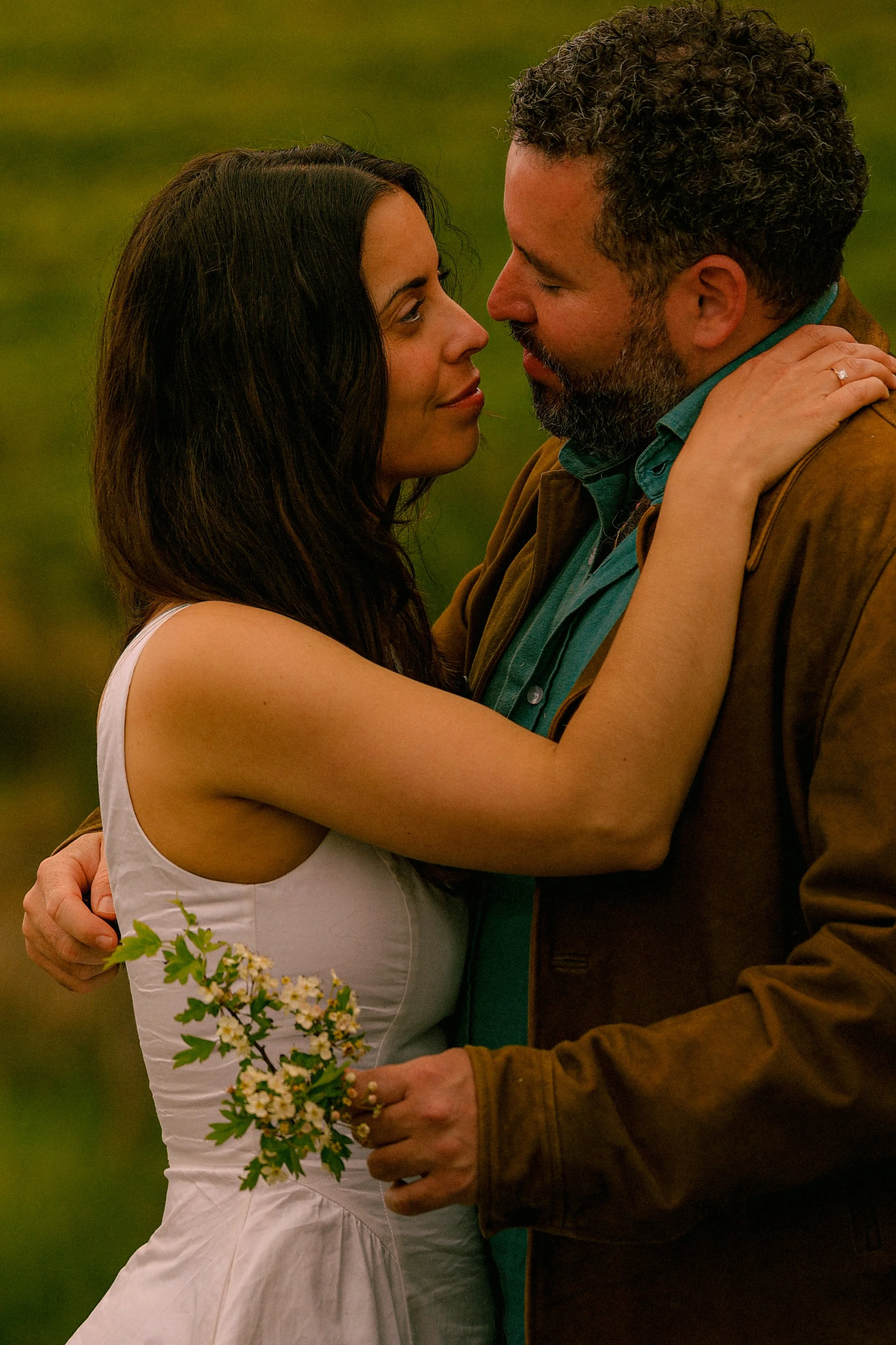 A man and woman holding each other closely outdoors, the woman holding a small bouquet of white flowers, both looking into each other's eyes.