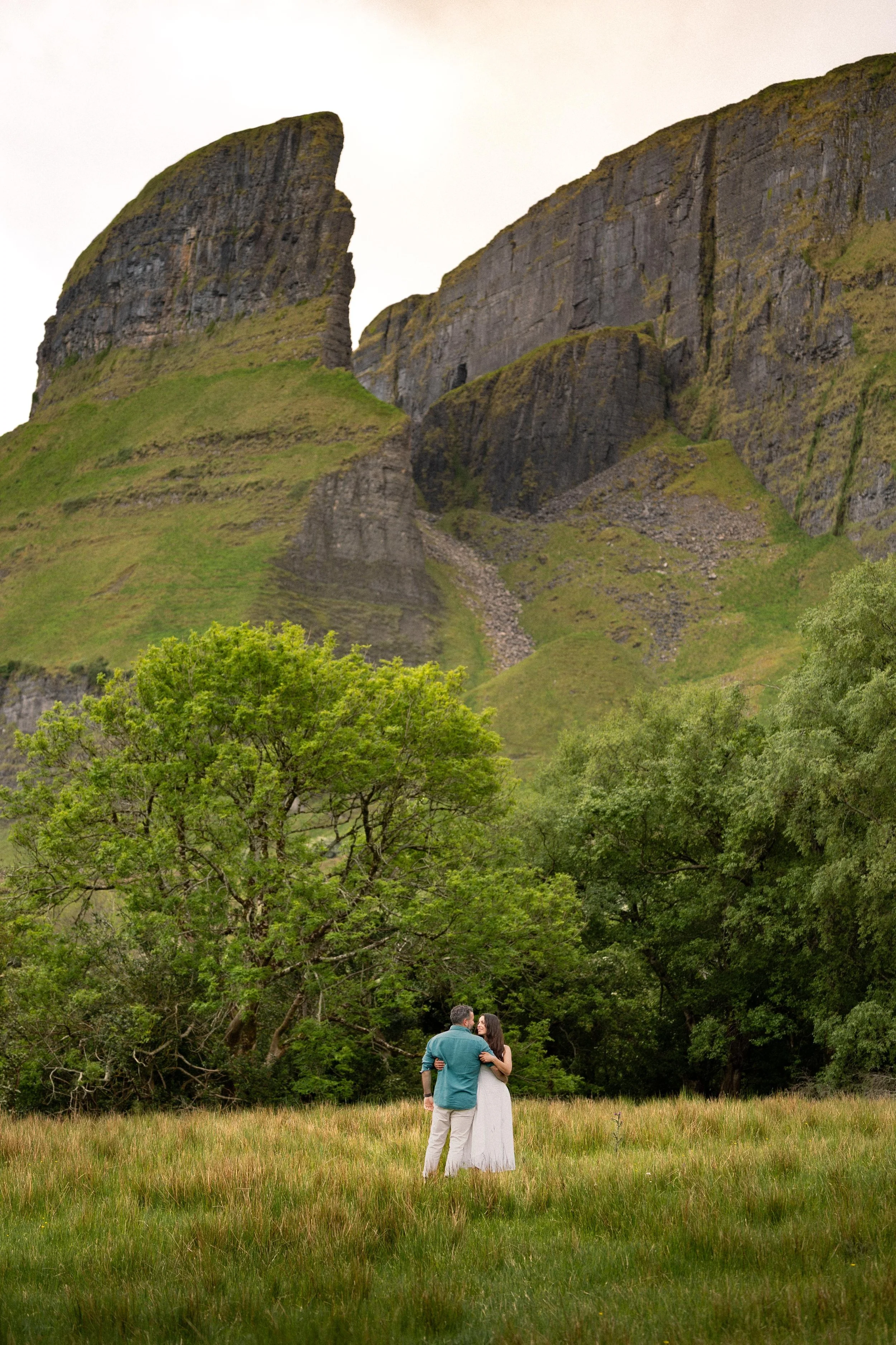 A couple standing in a grassy field surrounded by trees, with mountain cliffs in the background, embracing and looking at each other.