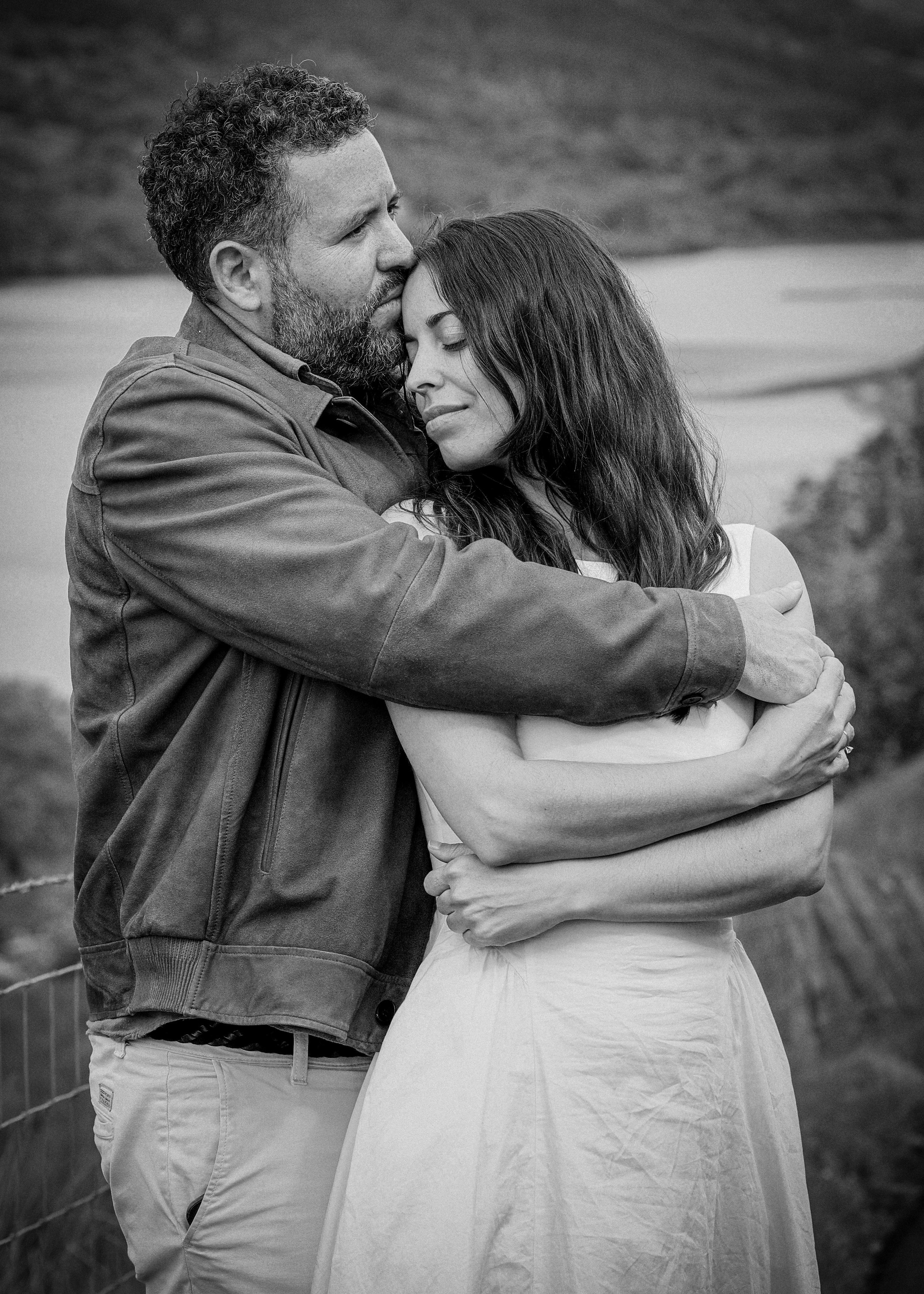 A black and white photo of a man with a beard and woman with long dark hair embracing outdoors. The man gently kisses the woman's forehead with eyes closed, and she has a serene expression with eyes closed, leaning into him.