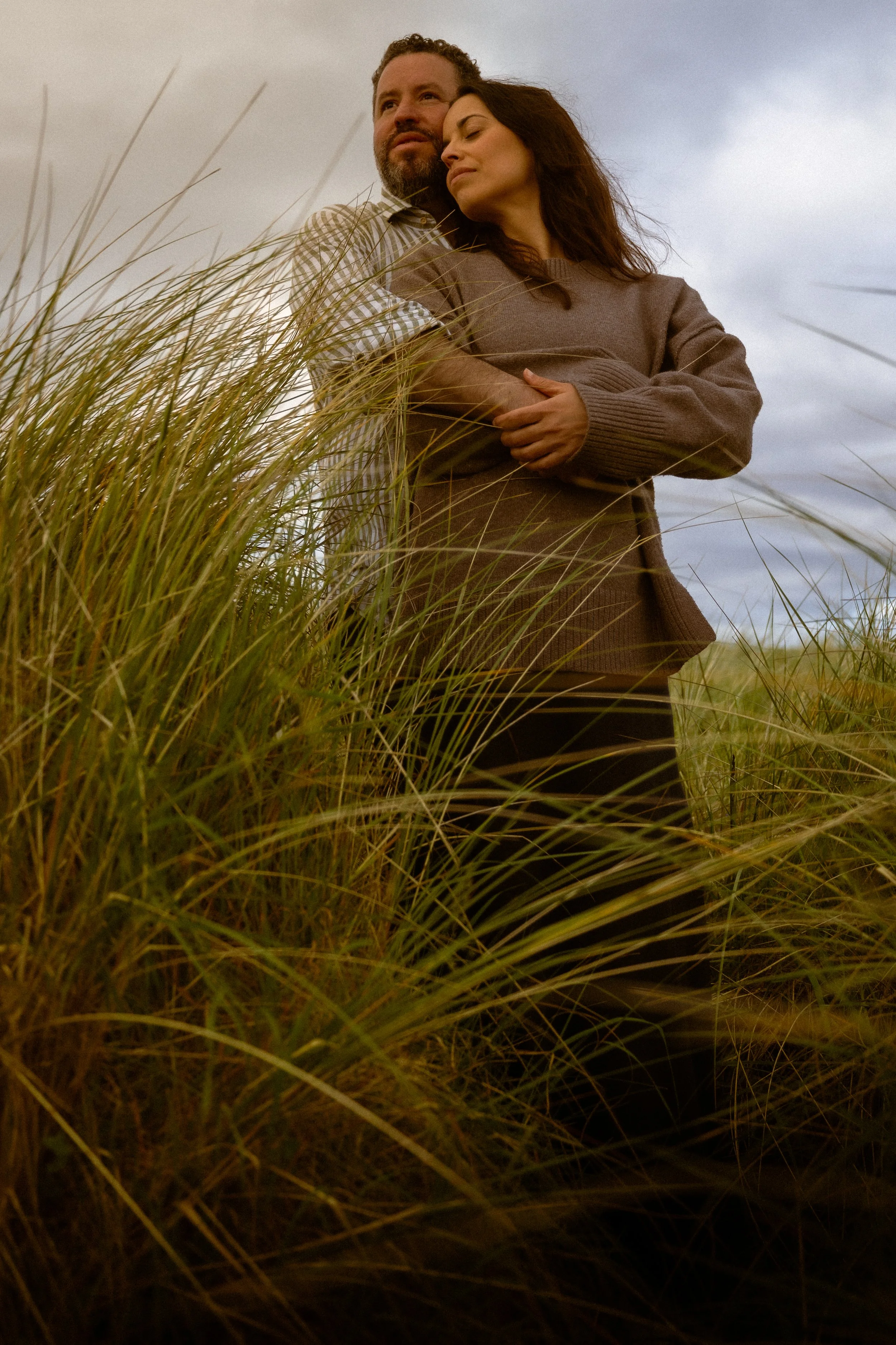 A man and a woman standing amidst tall grass outdoors, with cloudy sky in the background. The woman is resting her head on the man's shoulder with a peaceful expression, while the man looks off into the distance.
