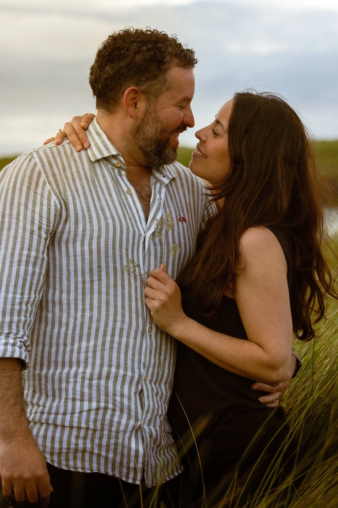 A happy couple stands close together in a field of tall grass, smiling and leaning in towards each other during sunset.