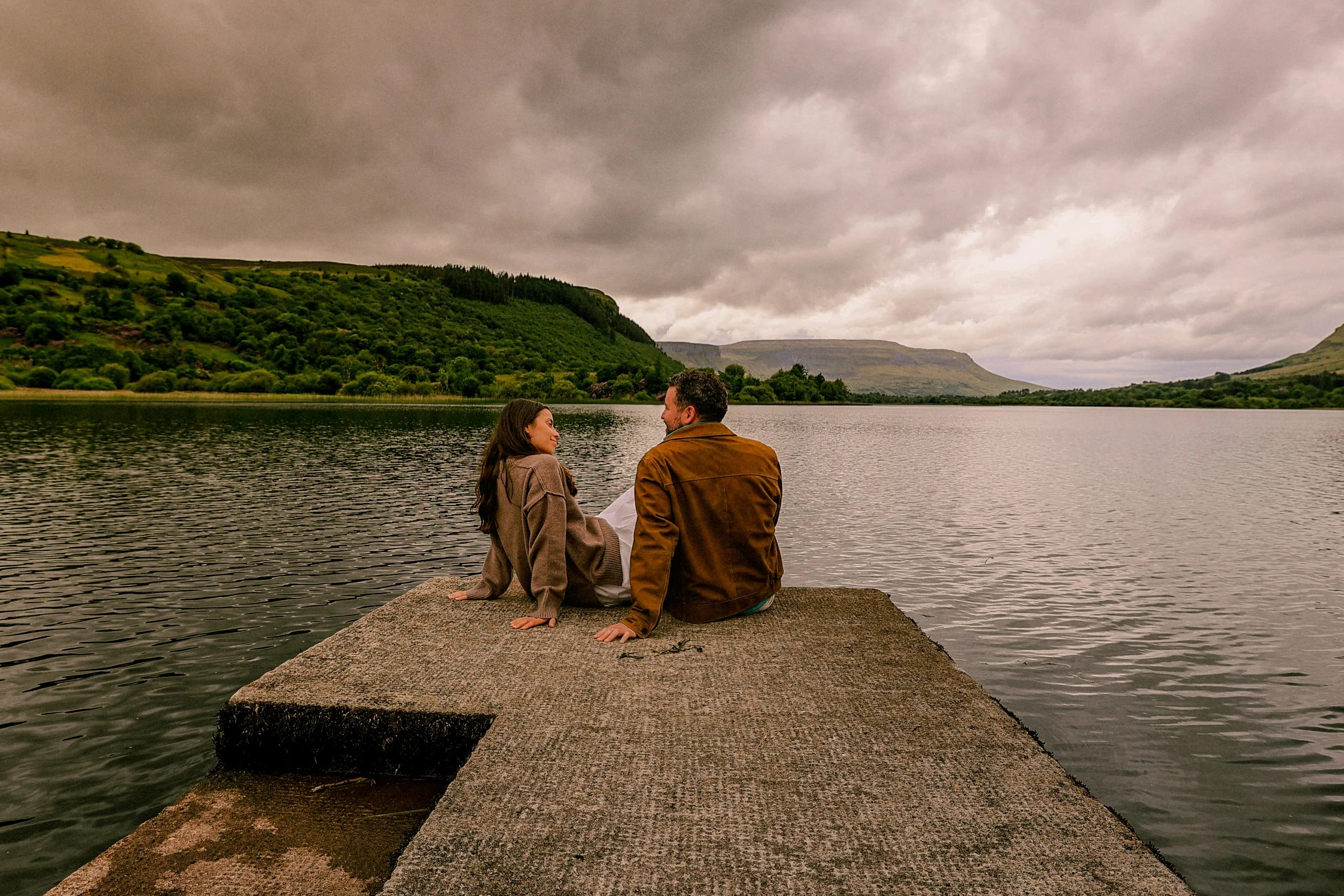 A man and a woman sitting on a stone dock overlooking a river, with green hills and cloudy sky in the background.