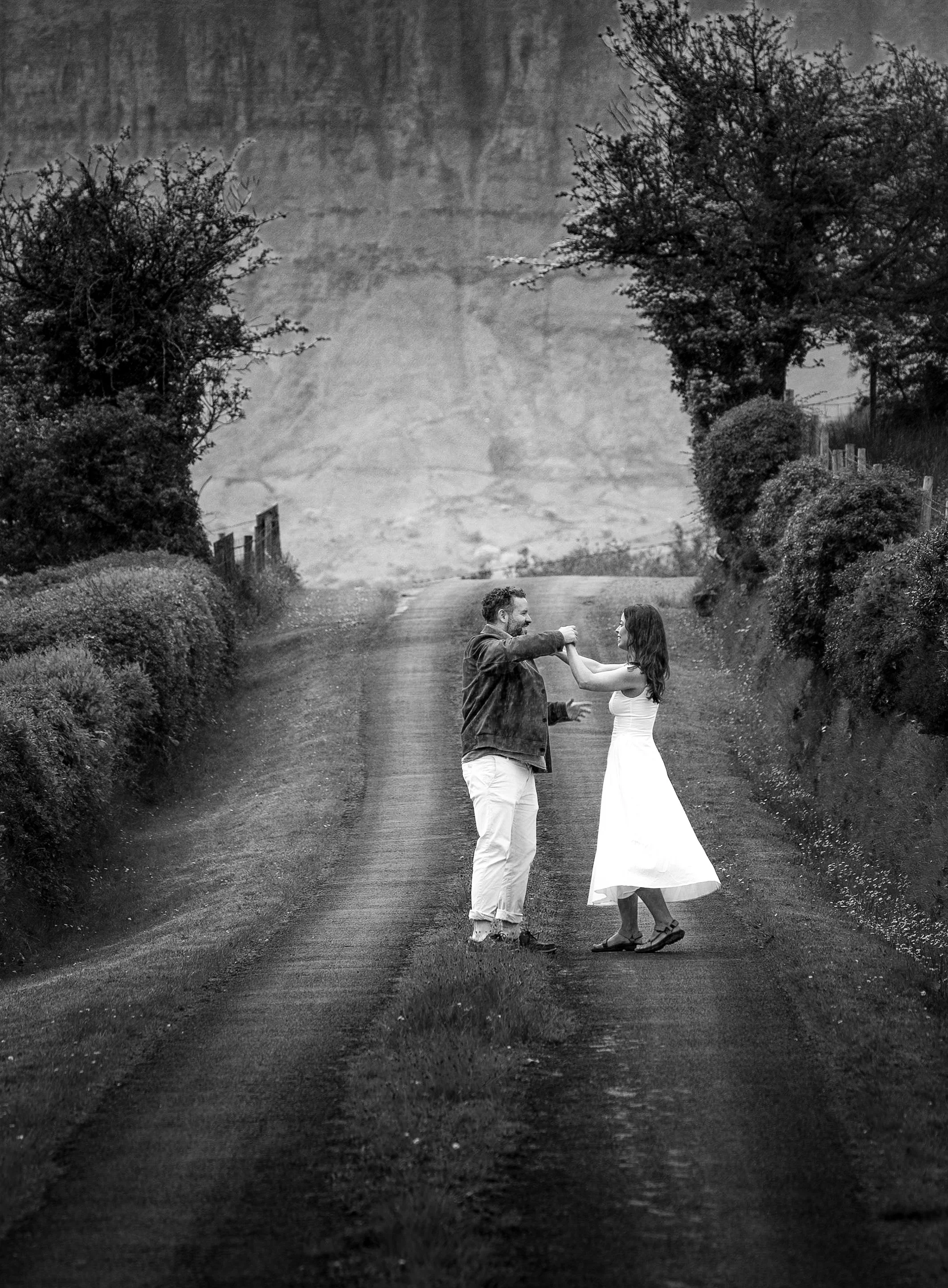 A black and white photo of a couple dancing in the middle of a rural dirt road, surrounded by bushes and trees, with hills in the background.