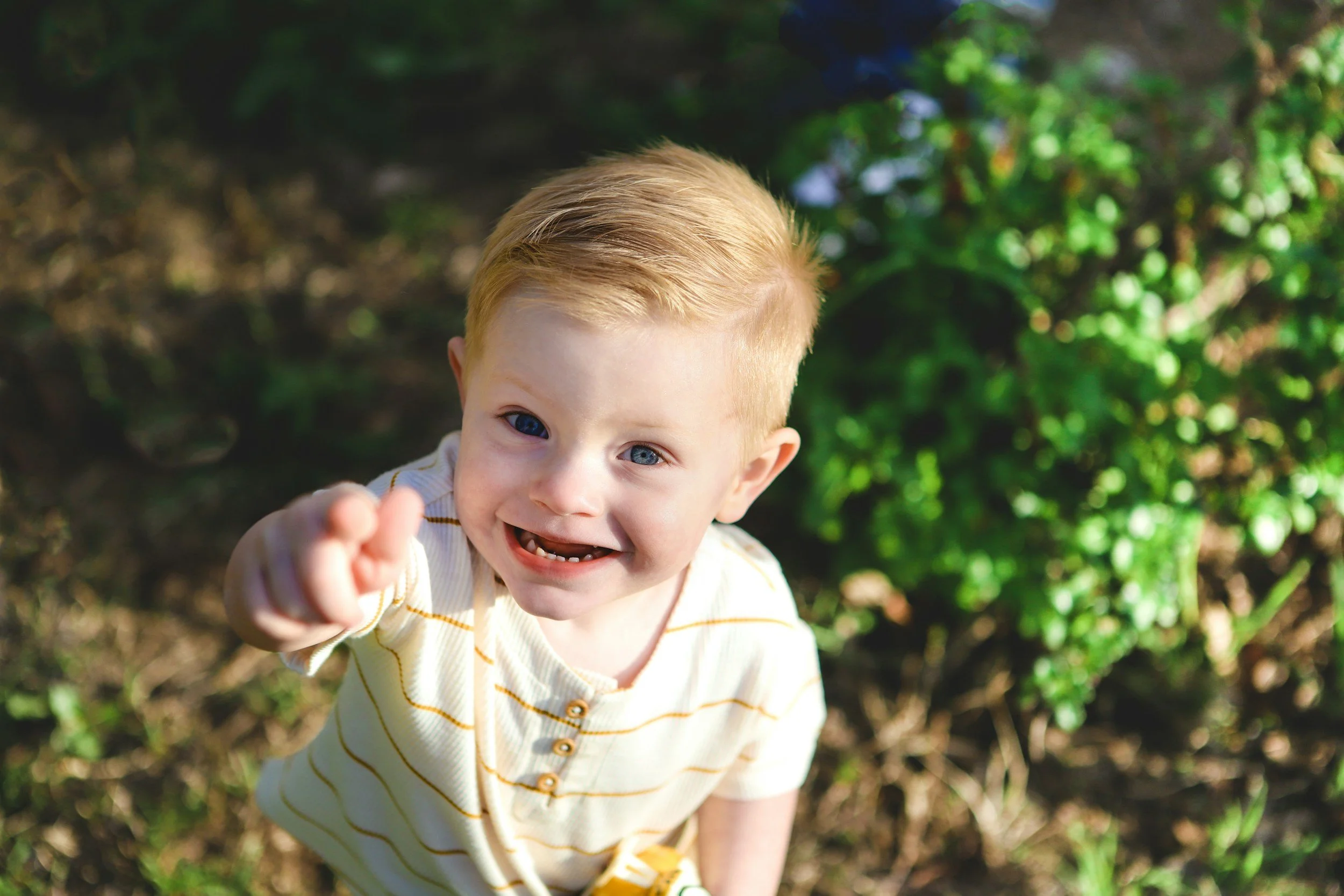 A young boy with blonde hair and blue eyes smiling and reaching out towards the camera outdoors with green foliage in the background.