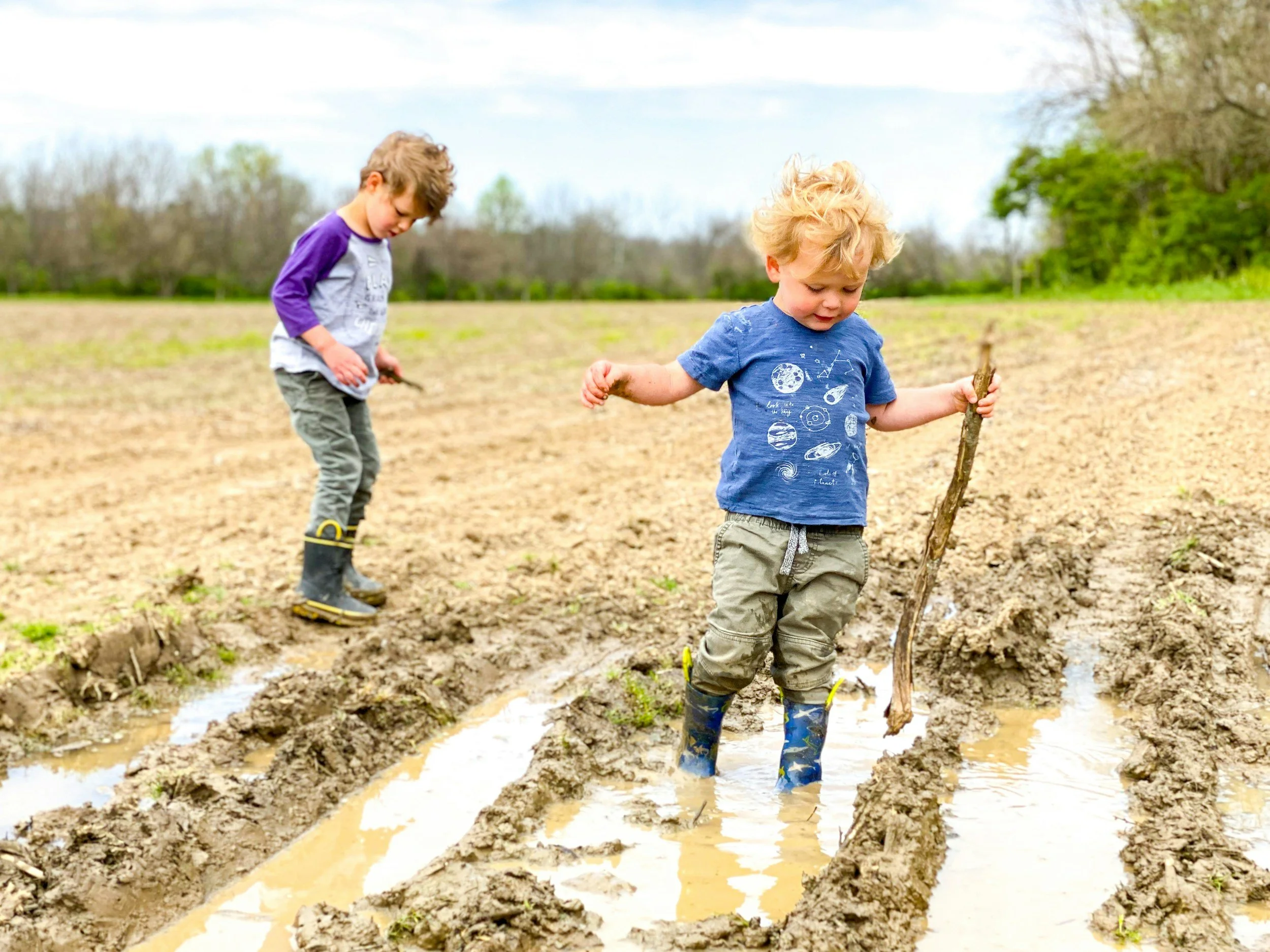 Two young children, a boy and a girl, are playing outdoors in a muddy field with water. The boy, wearing a blue shirt with space-themed graphics and rain boots, is holding a stick. The girl, wearing a purple and gray shirt and yellow rain boots, is standing nearby. The background shows trees and a cloudy sky.