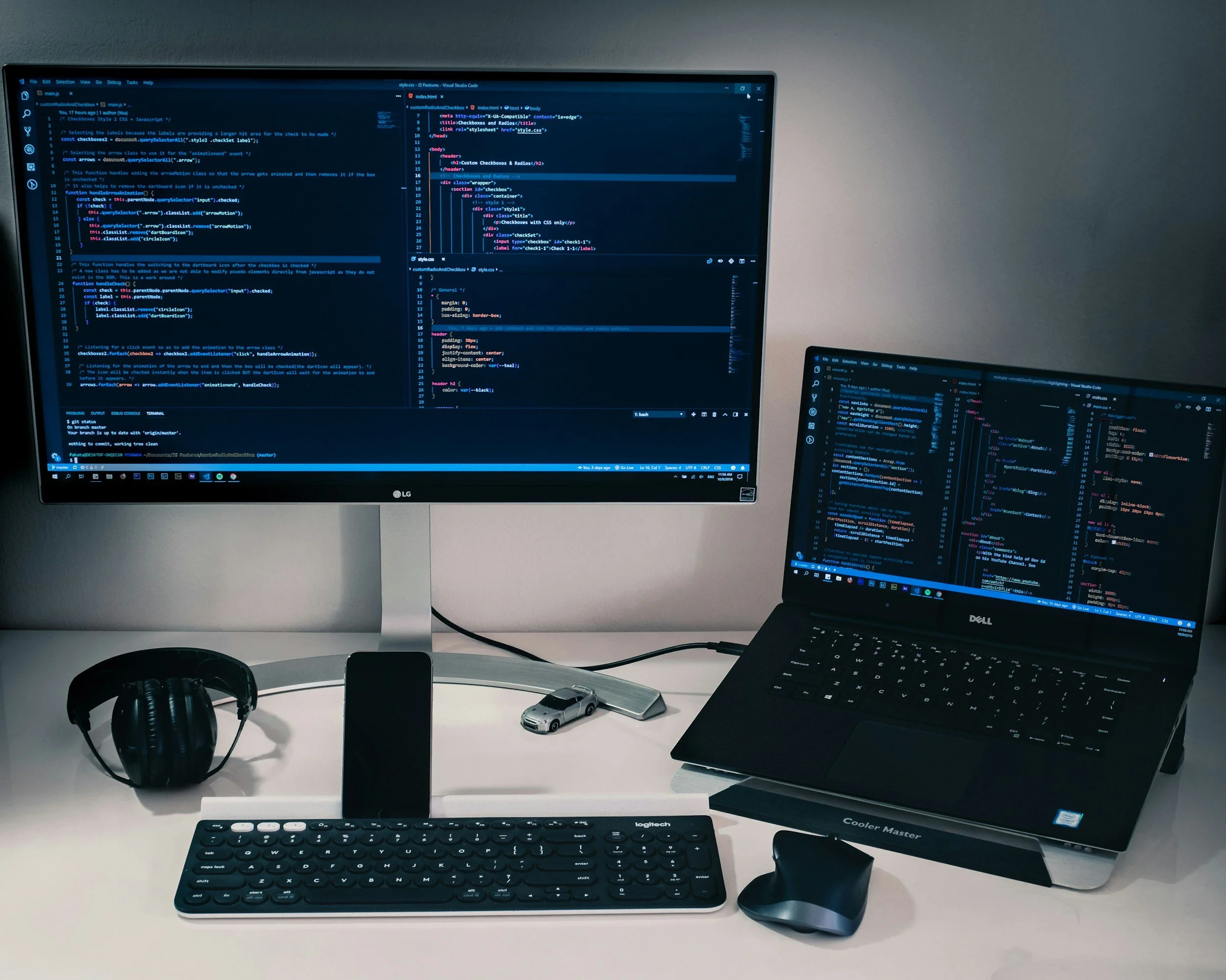 A computer desk setup with dual monitors displaying code, a keyboard, mouse, headphones, a phone, a small scale model car, and a tablet on a white surface.