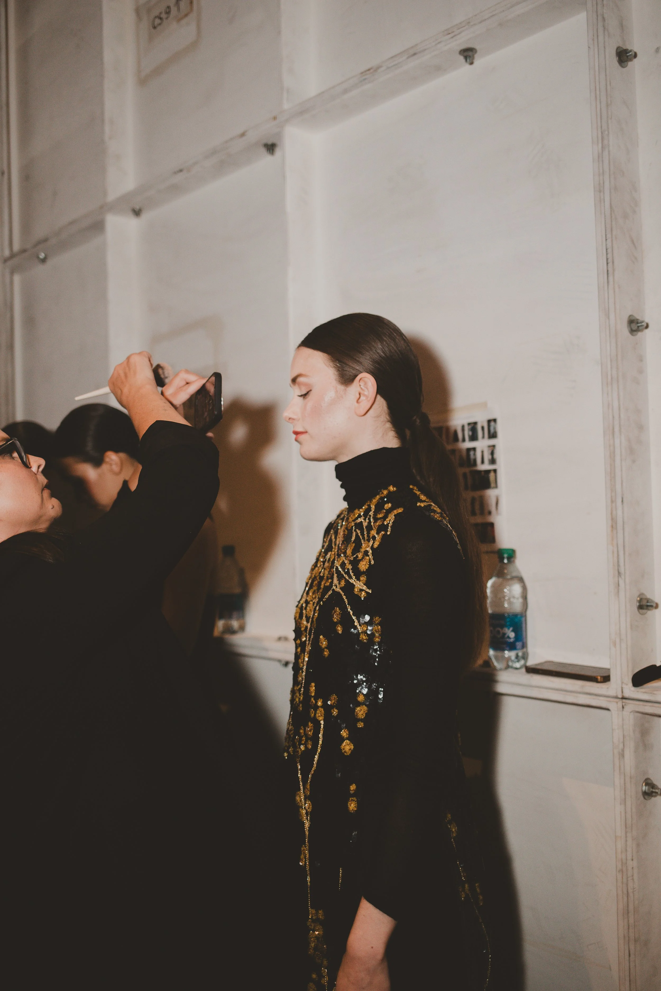 A woman with long dark hair styled back, wearing a black dress with gold embroidery and sequins, is having her makeup or hair done by a stylist at an indoor event. The stylist is holding a phone with one hand and a brush or makeup tool with the other. There is a water bottle on a table behind them, and a white wall with a photo collage and some hardware.