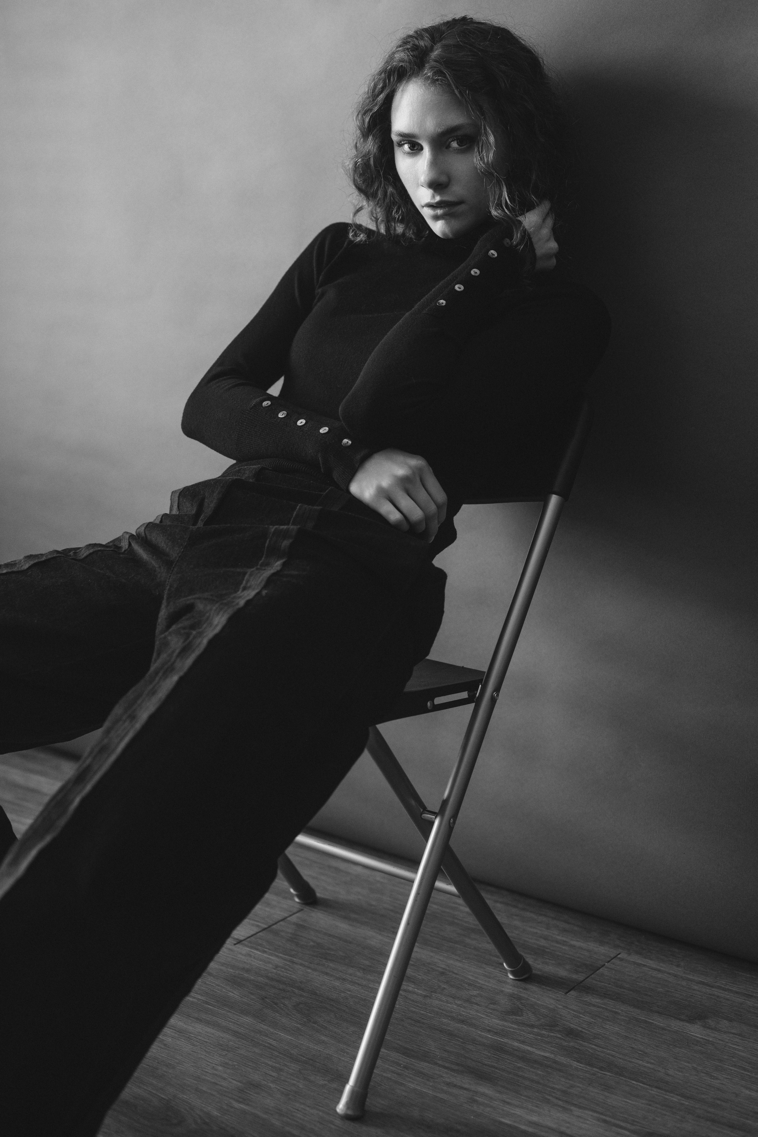A woman with curly hair sitting on a chair with her hand resting on her neck, wearing dark clothing, in a black and white photo.