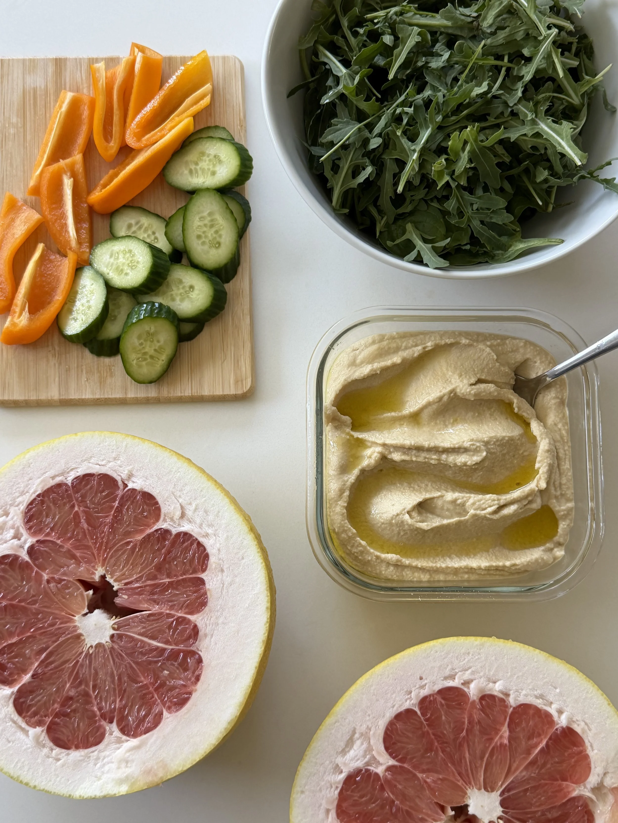 Slices of pink grapefruit, a cucumber, sliced yellow bell peppers, fresh arugula in a bowl, and hummus with a drizzle of olive oil.