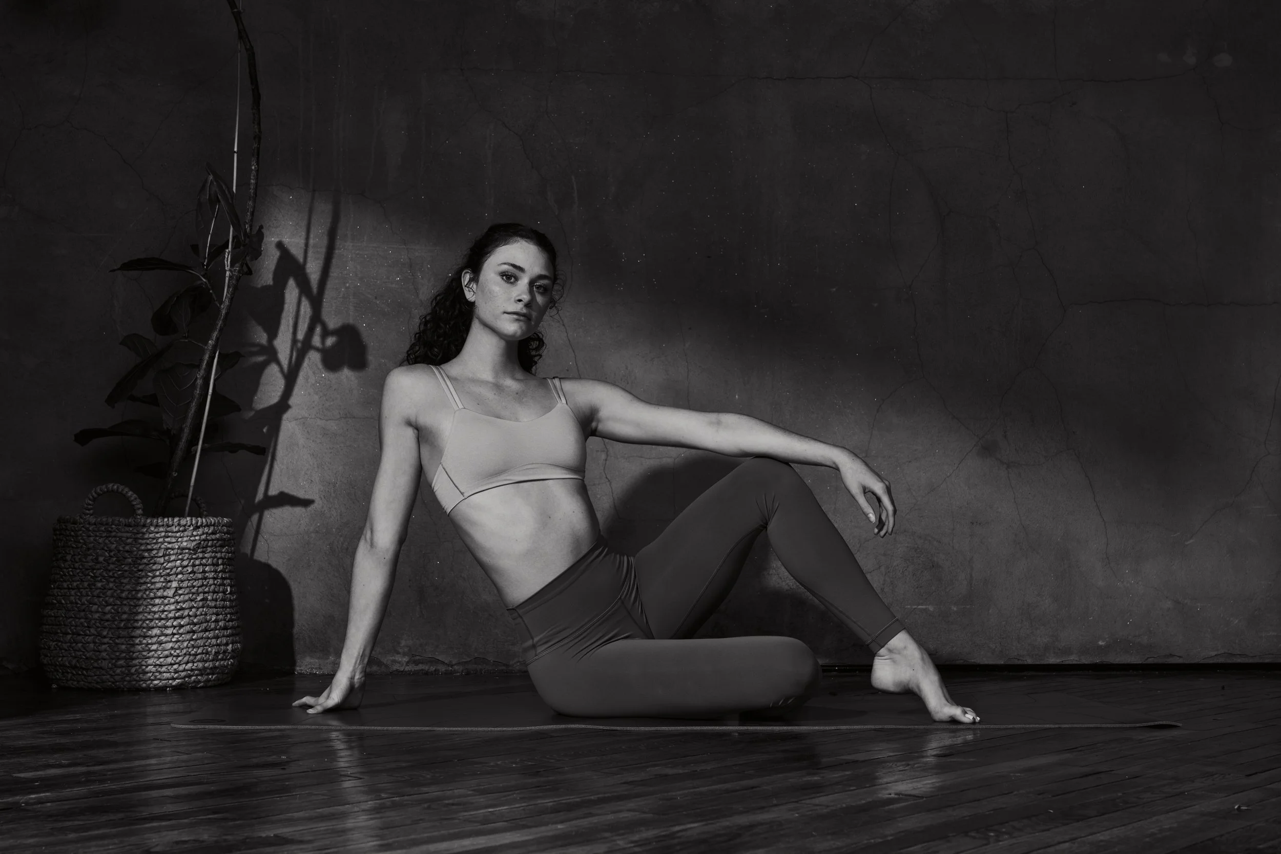 A woman in athletic wear performs a yoga pose on a yoga mat indoors, with a large potted plant beside her, against a textured dark wall.