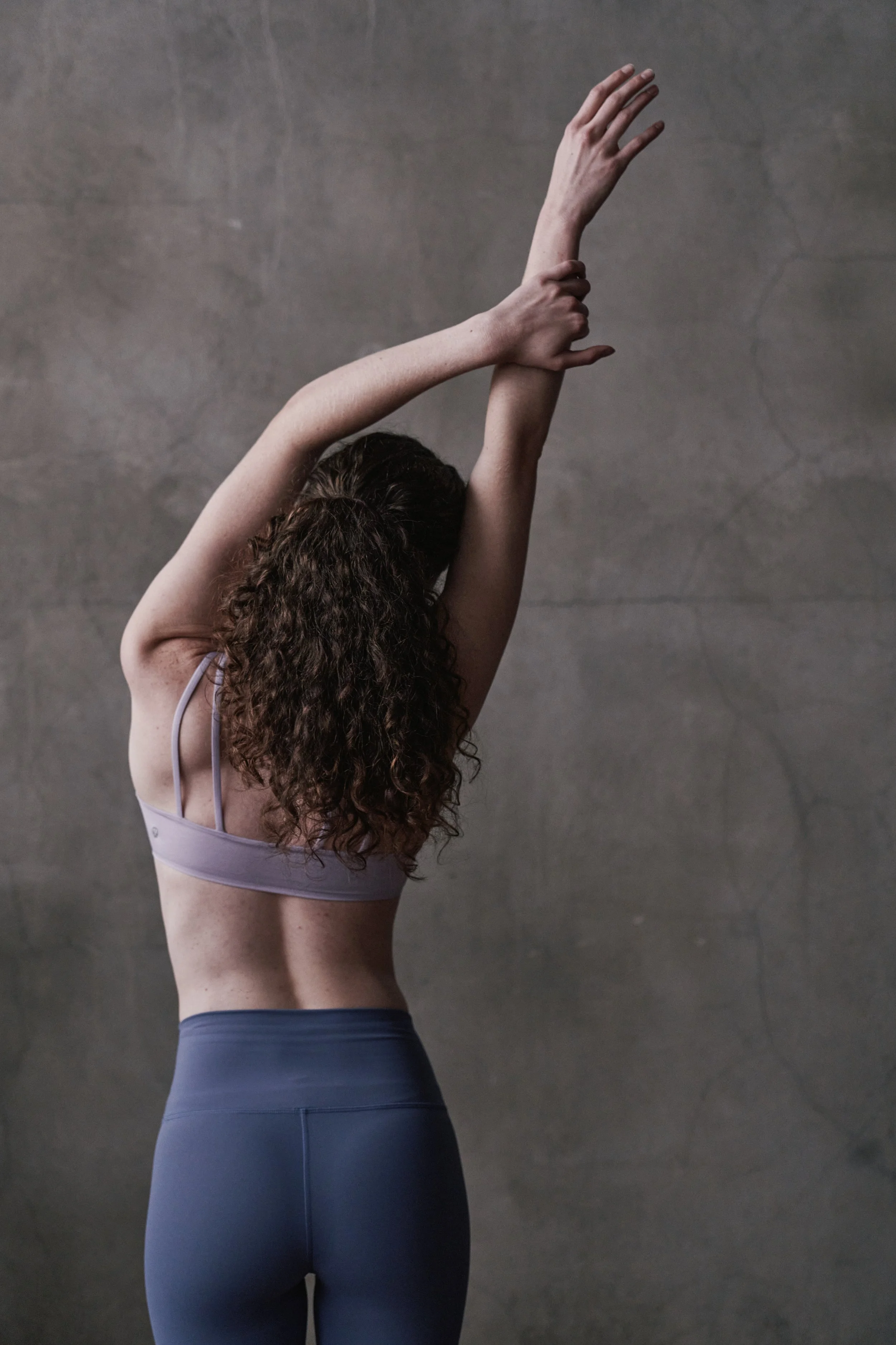 A woman with curly hair in a workout bra and leggings stretching her arms overhead against a plain gray wall.