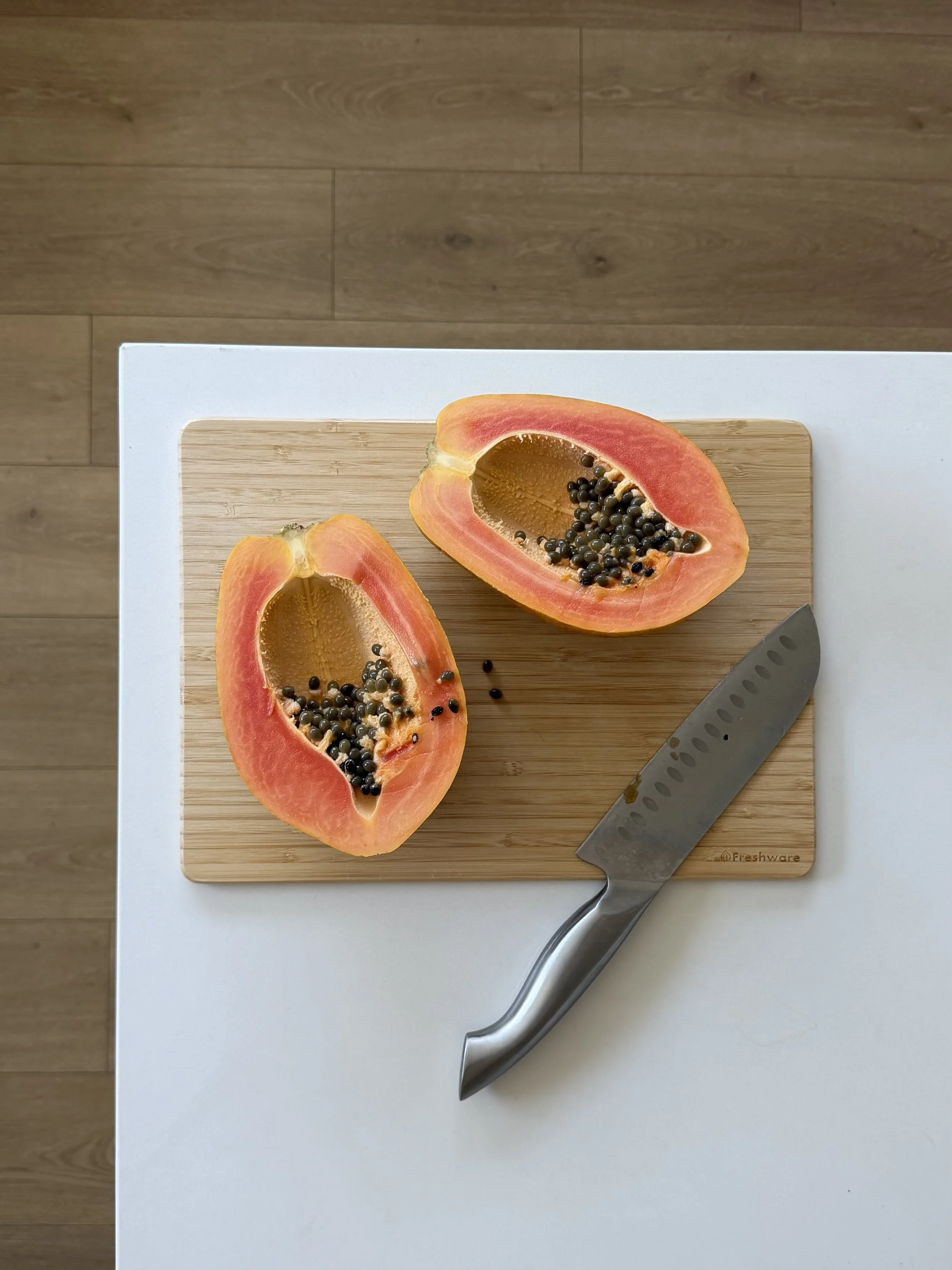 A halved papaya on a wooden cutting board with a stainless steel knife beside it, resting on a white surface.