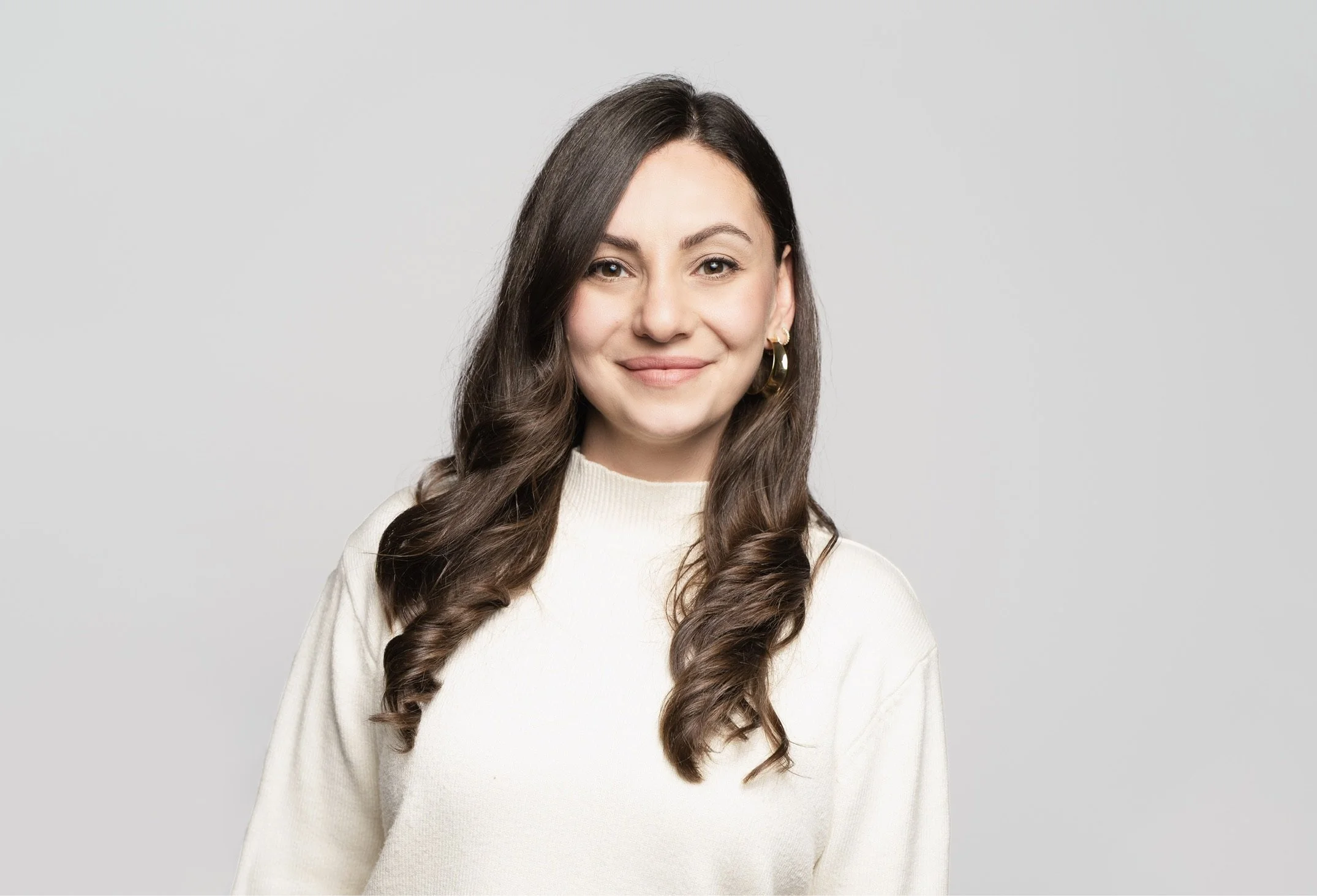 A woman with long, wavy brown hair, wearing a cream-colored sweater, smiling softly against a plain light gray background.