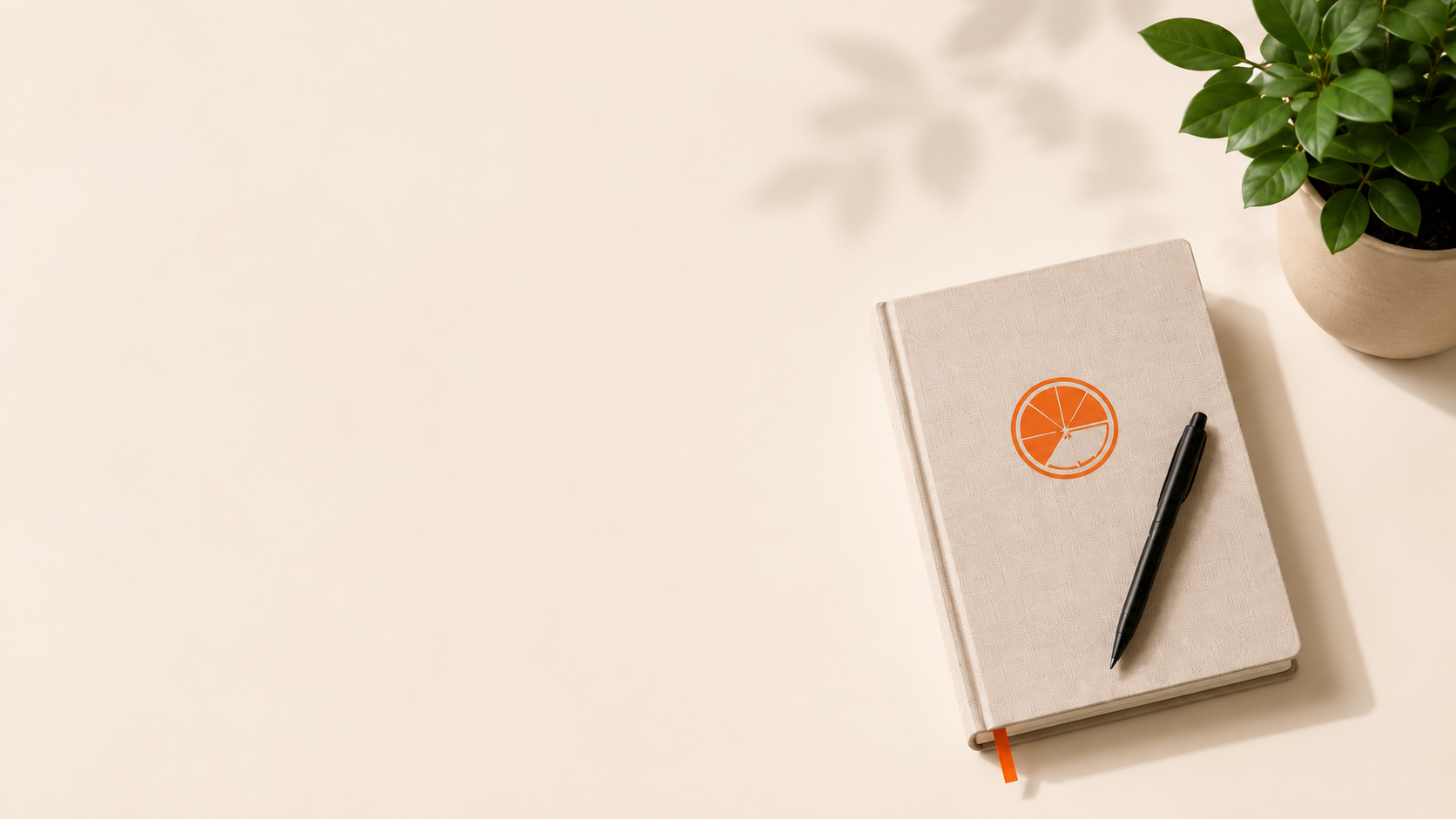 A beige notebook with an orange citrus slice graphic on the cover, a black pen resting on it, and a potted green plant on a white surface.