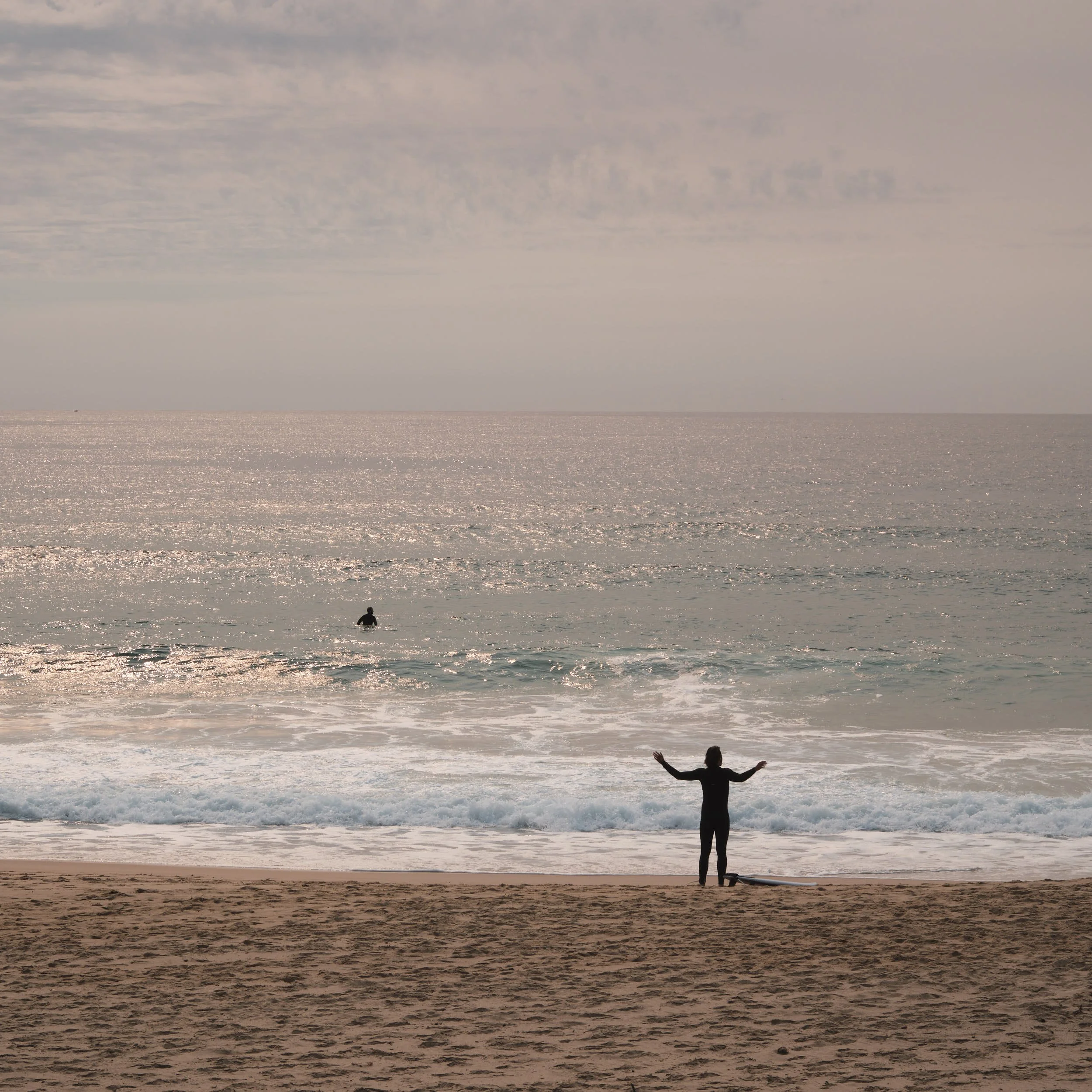 Bild zeigt eine Person im Wasser und eine andere Person am Strand. Neben ihr ein Surfboard.