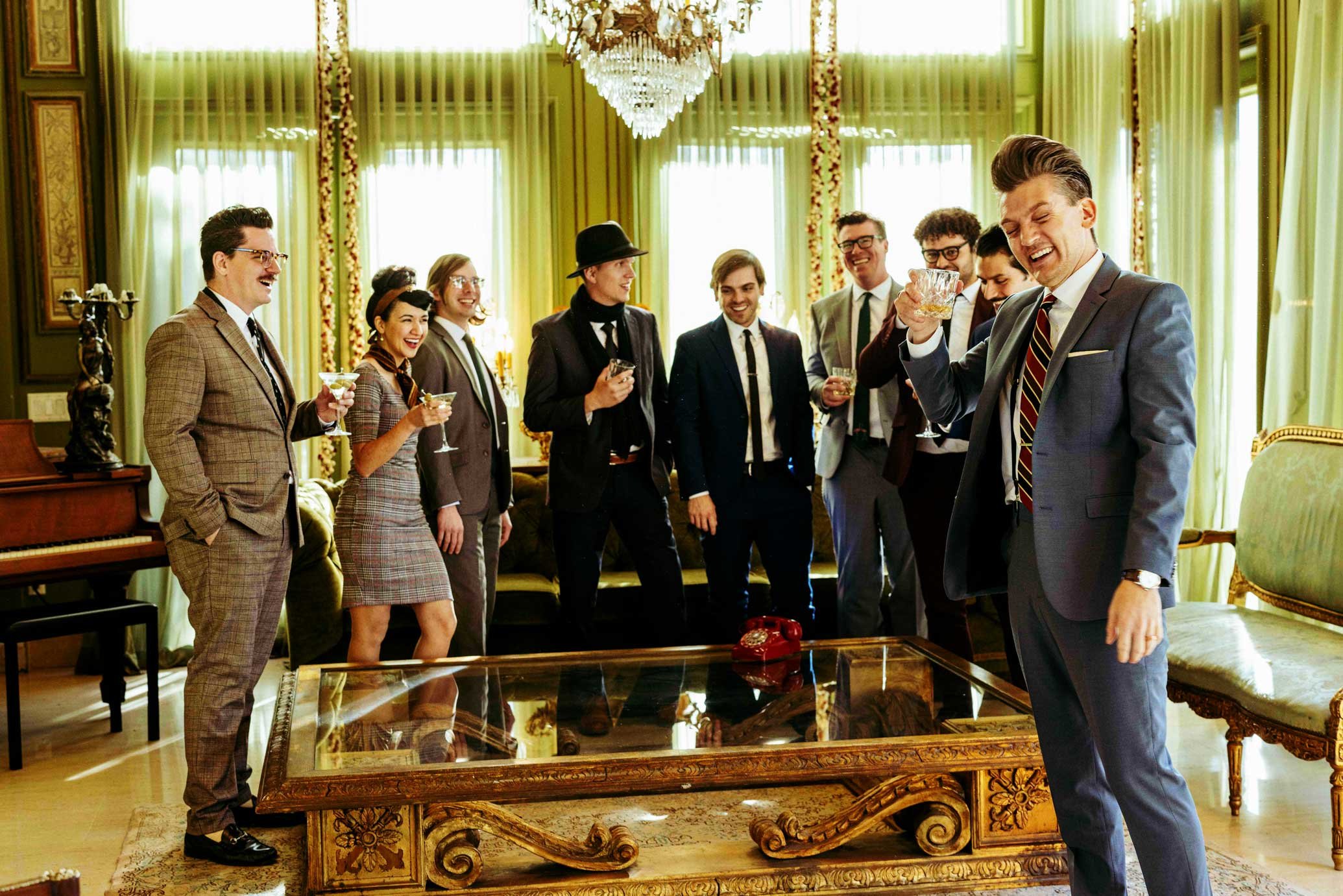 A group of people dressed in vintage style suits and dresses are gathered in an elegant room with green curtains, a chandelier, and ornate furniture. They are smiling and raising glasses, toasting at a celebration.