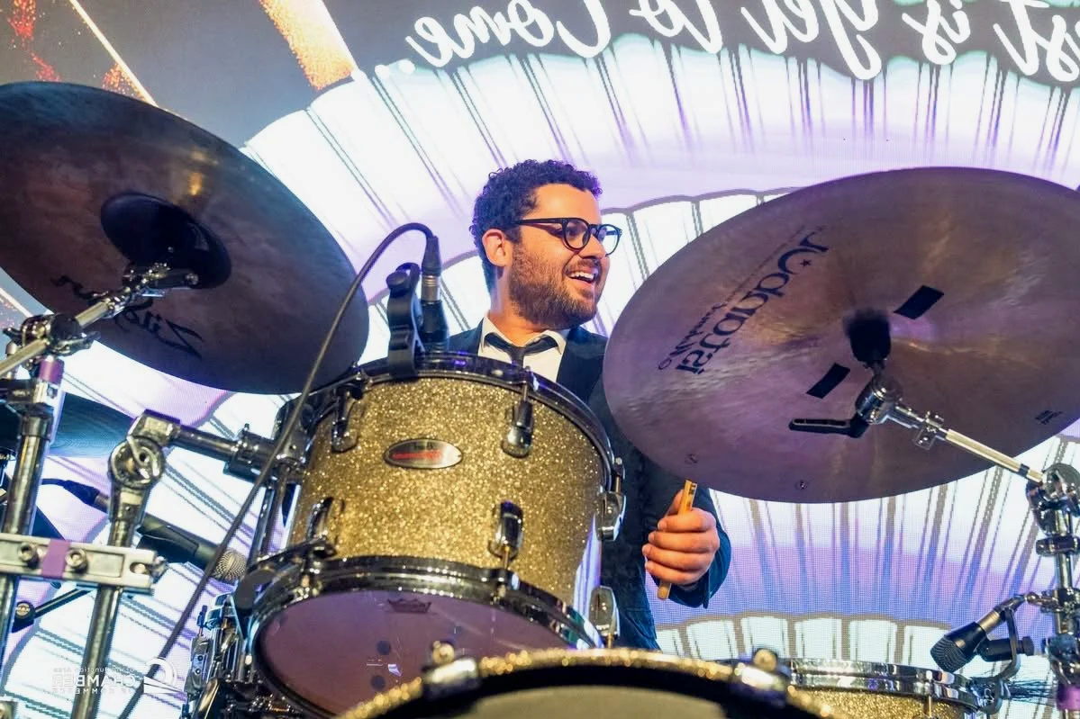 A man with glasses on a drum set, smiling, at a concert, with a colorful light show behind him.