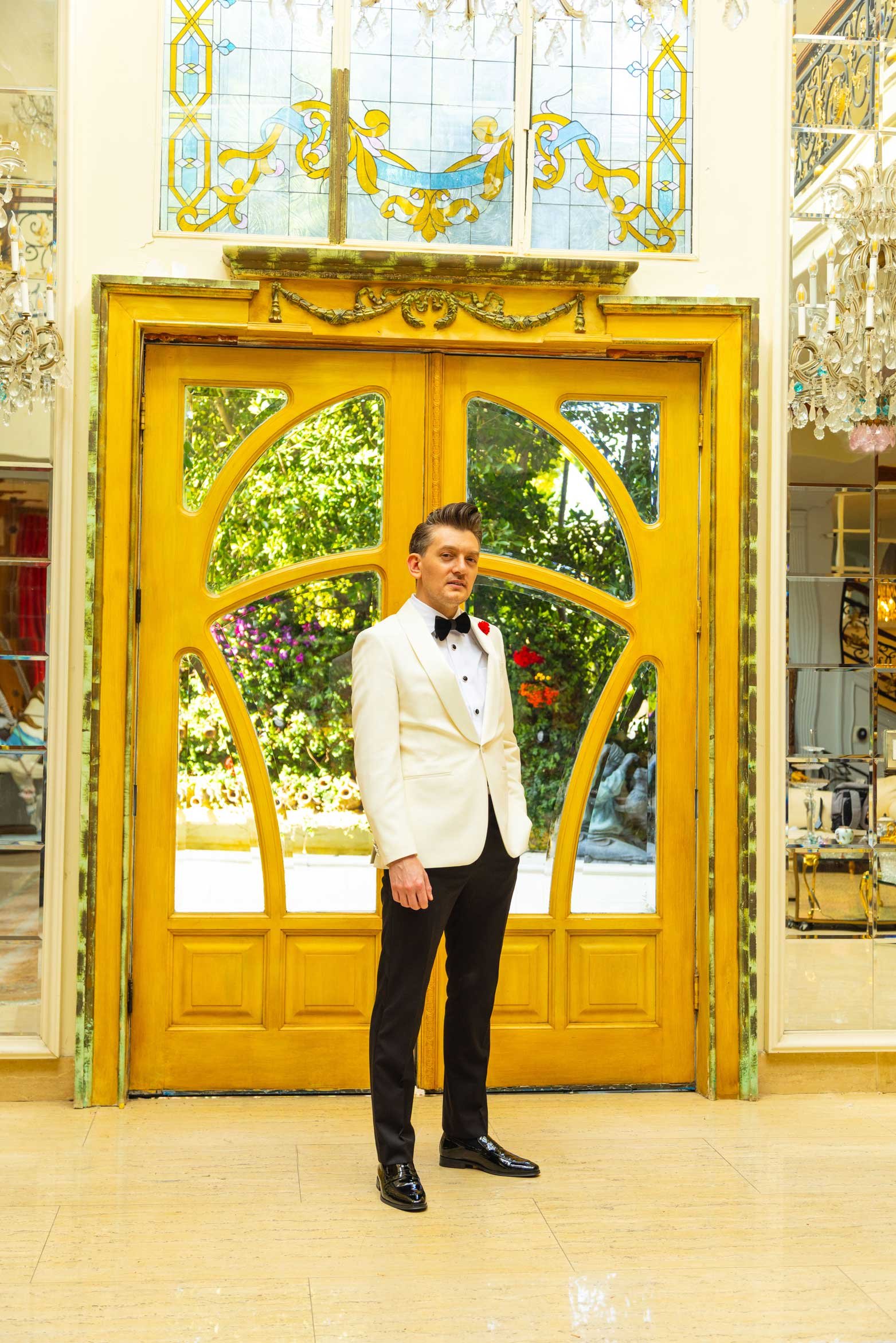 Man in a white tuxedo jacket, black bow tie, and black pants standing in front of a yellow wooden door with glass panels, decorated with greenery and flowers visible through the glass.