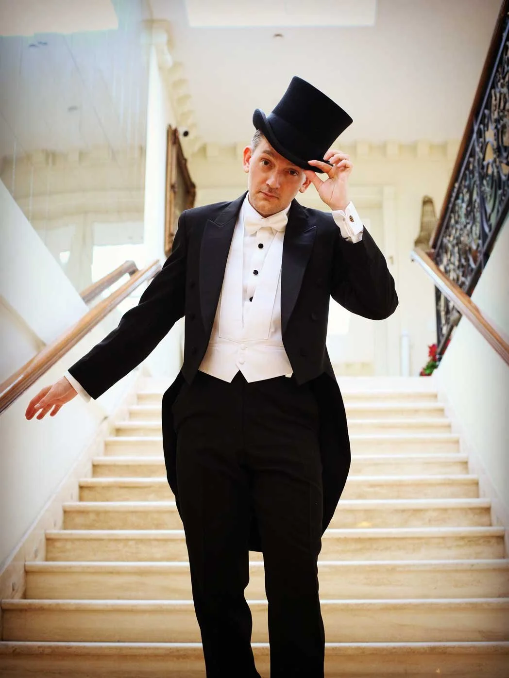 A man dressed in formal attire with a tuxedo, white shirt, and black bow tie, holding a black top hat and standing on a staircase indoors.