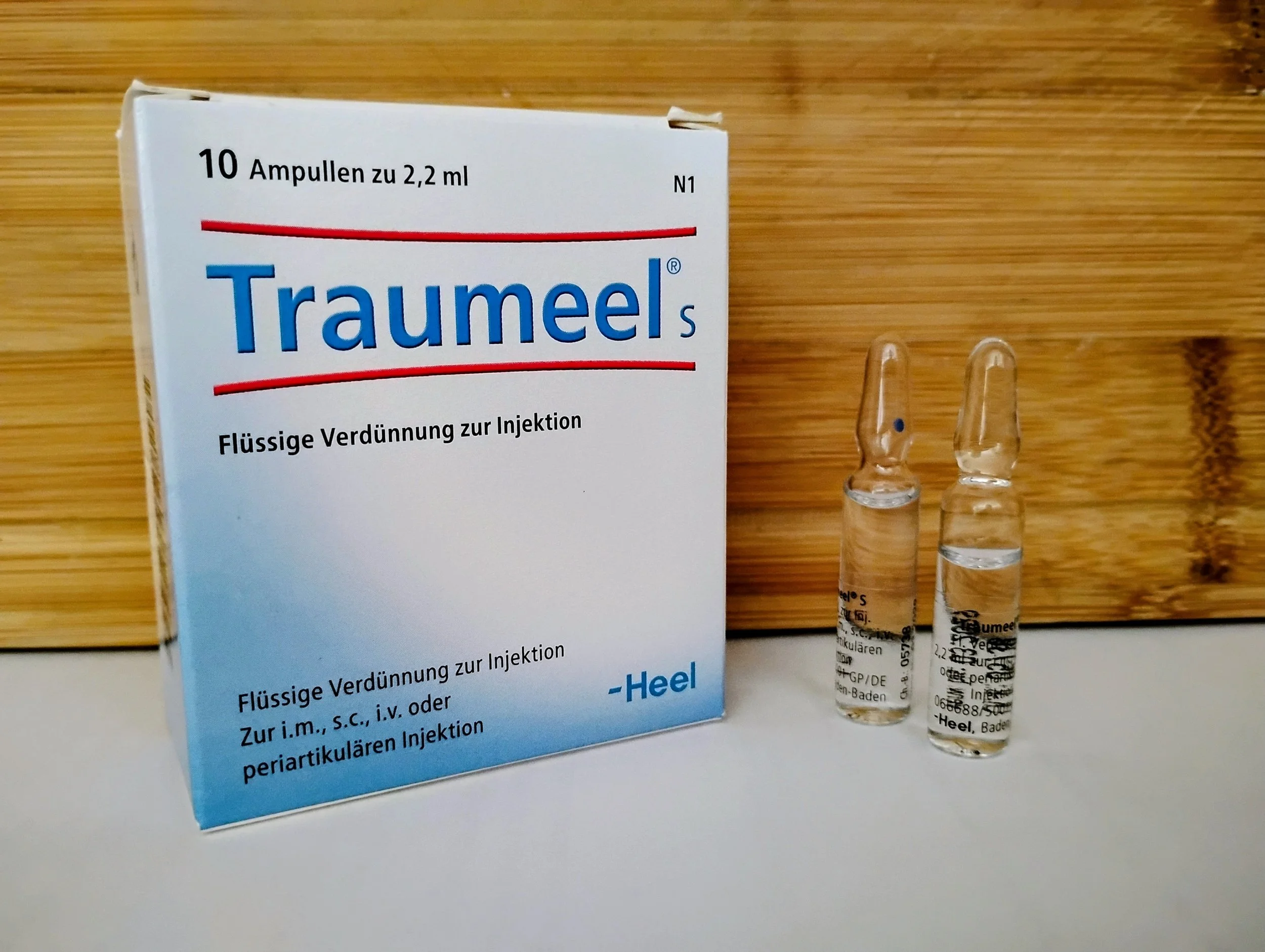Box of Traumeel ampoules with two ampoules standing upright next to it, on a wooden surface.