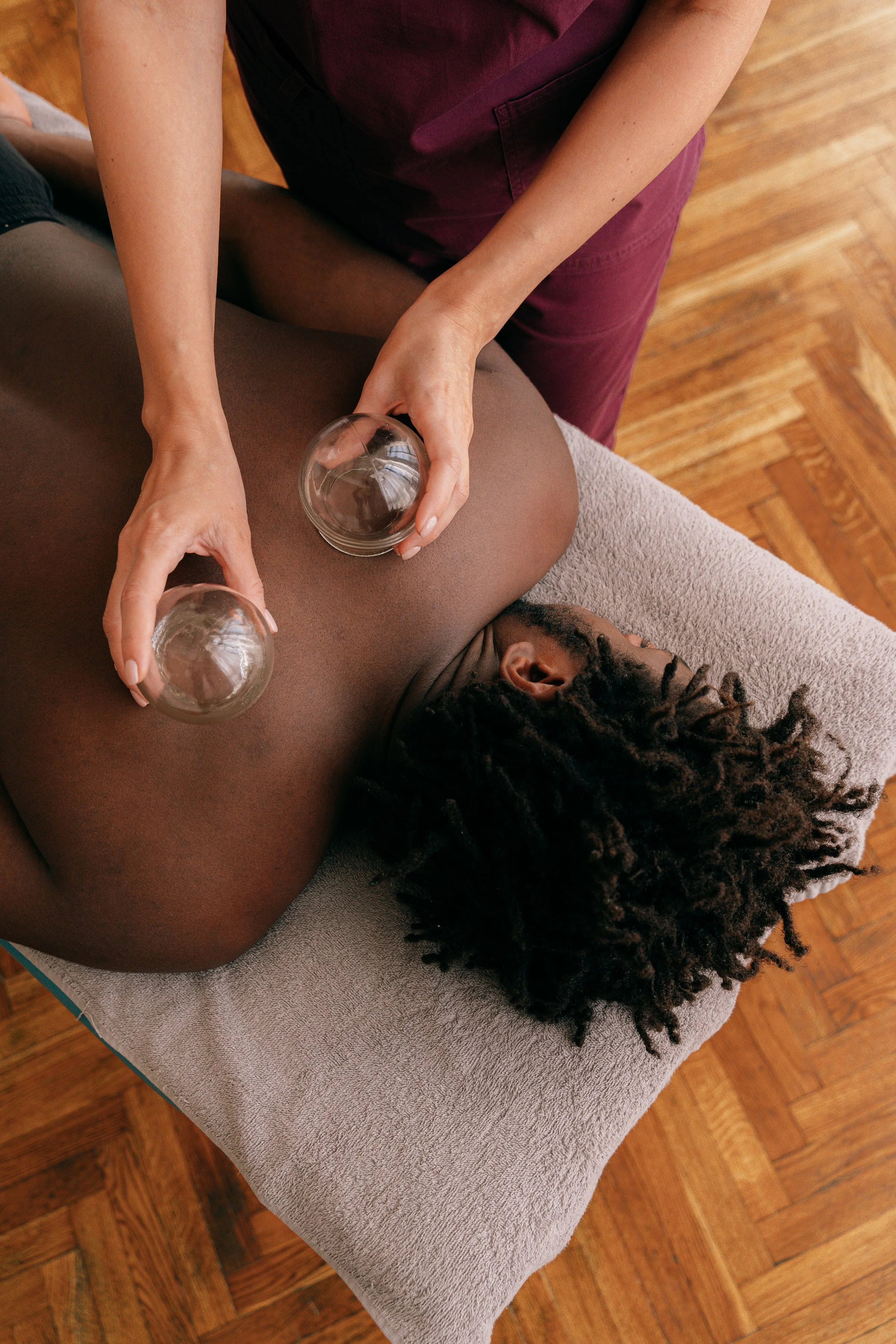 A person lying face down on a massage table, receiving a massage while holding two glass bowls filled with water. The setting appears to be a wellness or massage therapy room with wooden flooring.
