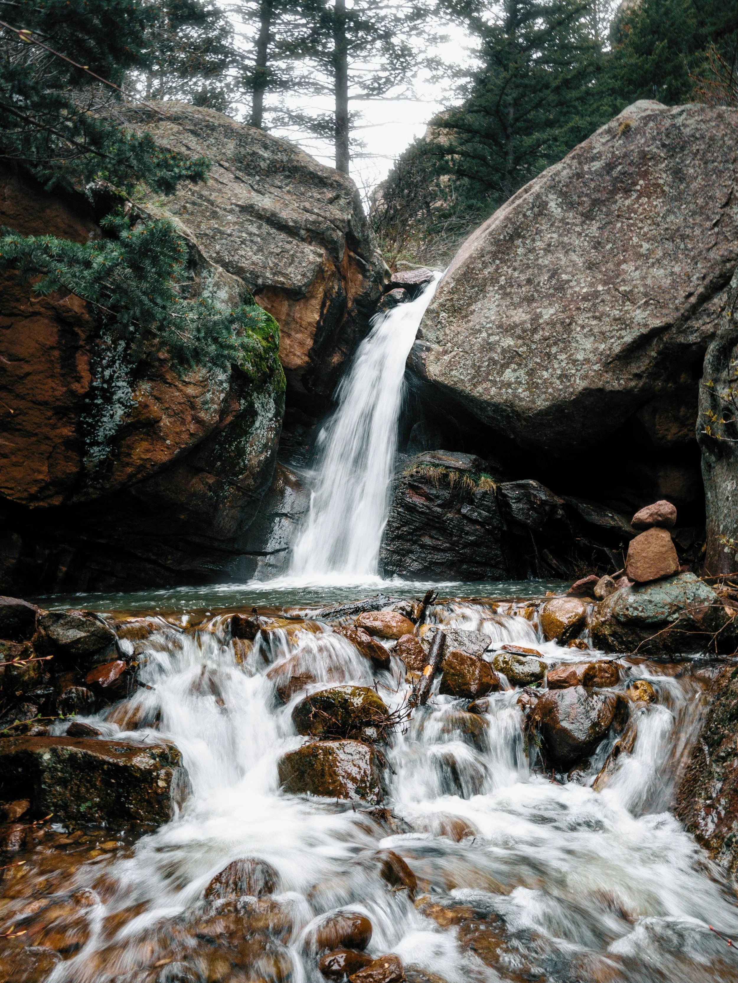 A small waterfall flowing between large rocky boulders in a forest with evergreen trees in the background.