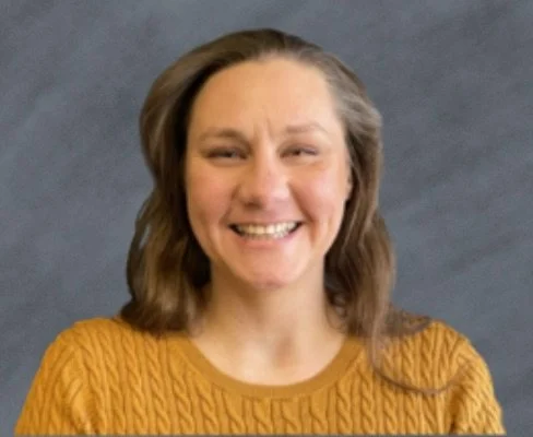 A woman with shoulder-length brown hair, smiling, wearing a mustard yellow cable knit sweater, standing against a dark gray background.
