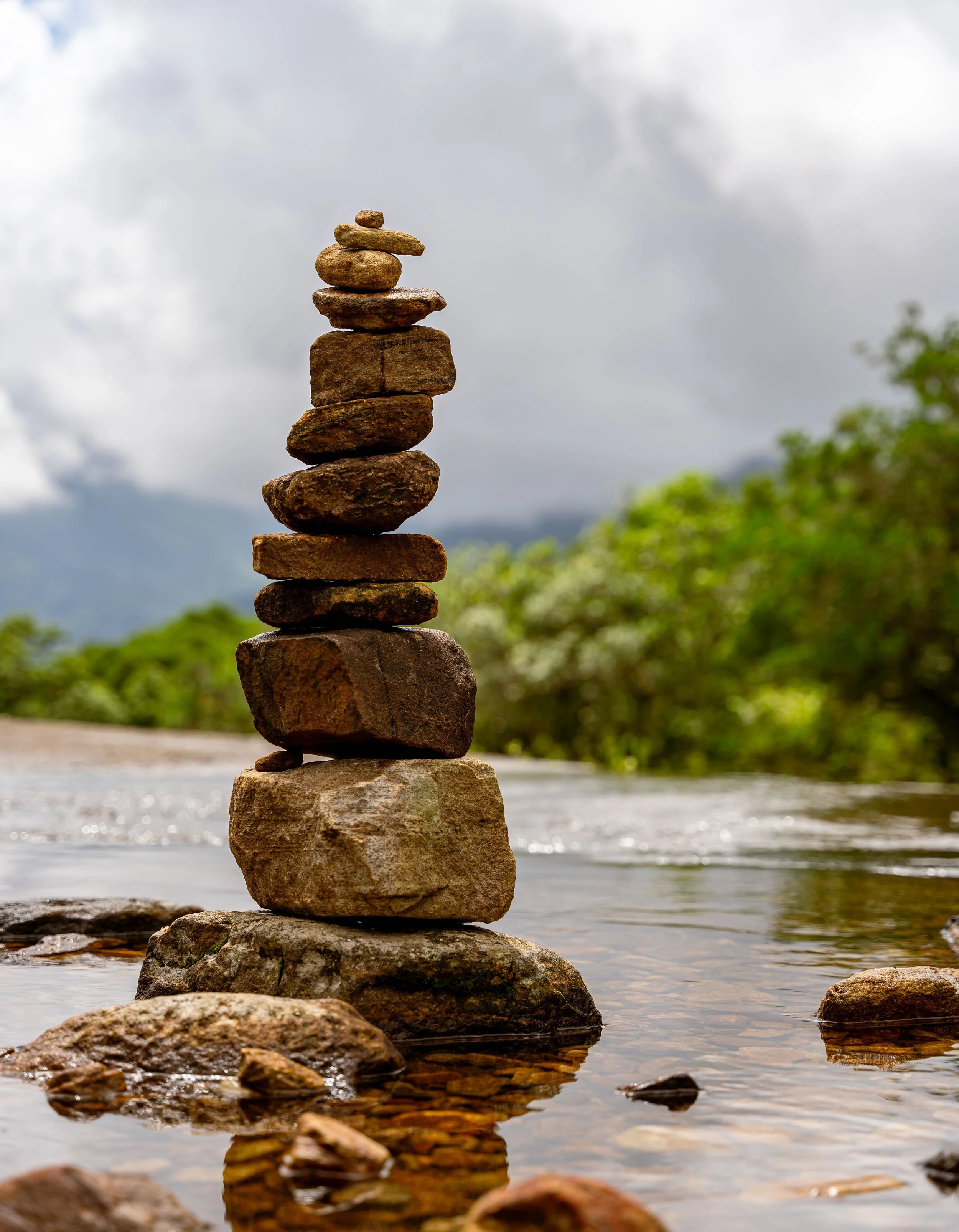 A stack of eleven rocks balanced on top of each other on a rock in a river, with a cloudy sky and greenery in the background.