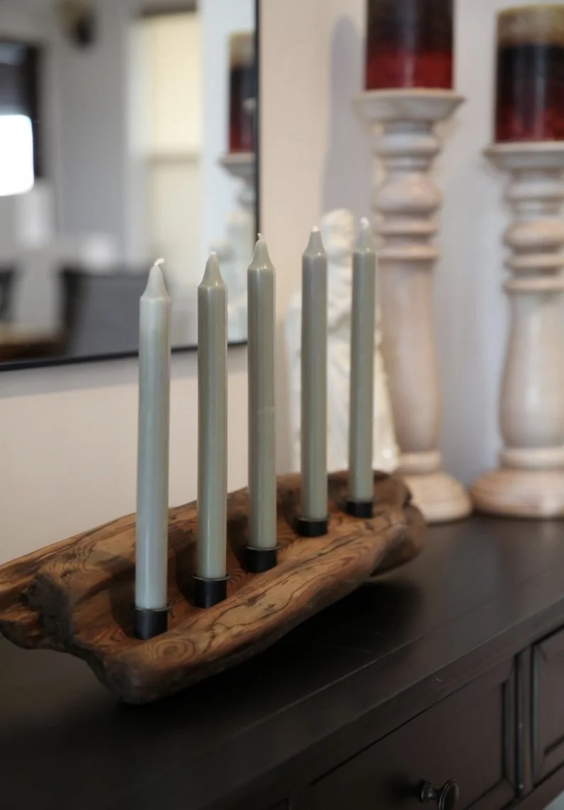 Decorative wooden candle holder with five white candles on a black surface, with large white candlesticks in the background.