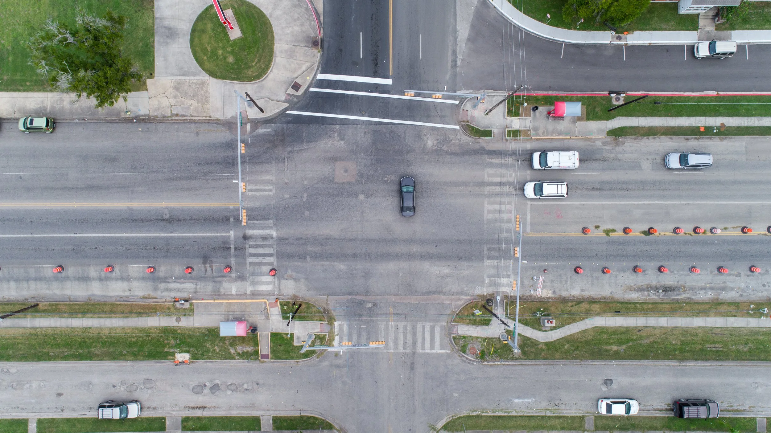 An aerial view of a city street intersection with cars, traffic cones, and sidewalks, showing an under-construction or roadwork area.