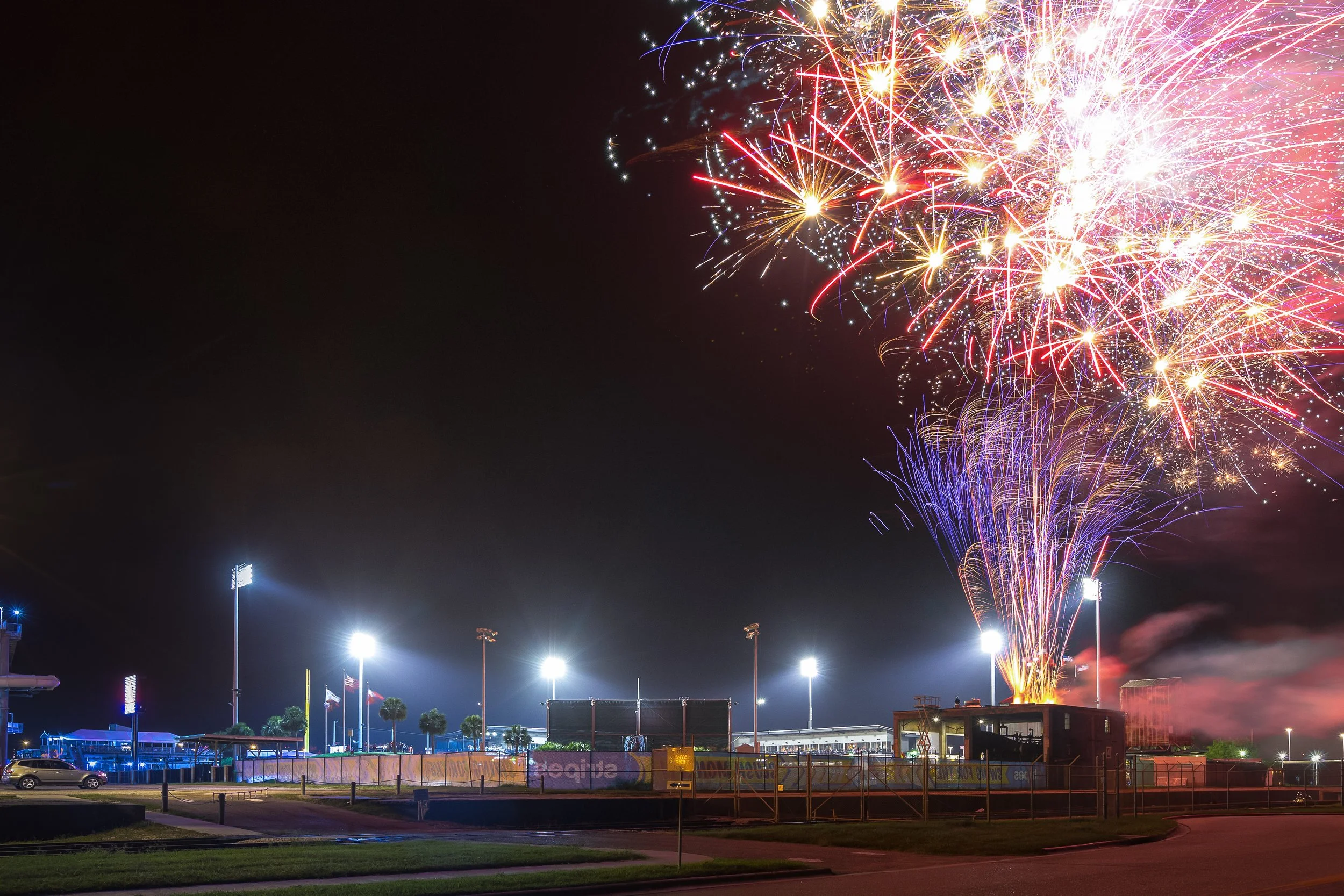 Fireworks explode above a stadium at night, illuminating the dark sky with colorful streaks, with stadium lights and parked cars visible in the foreground.