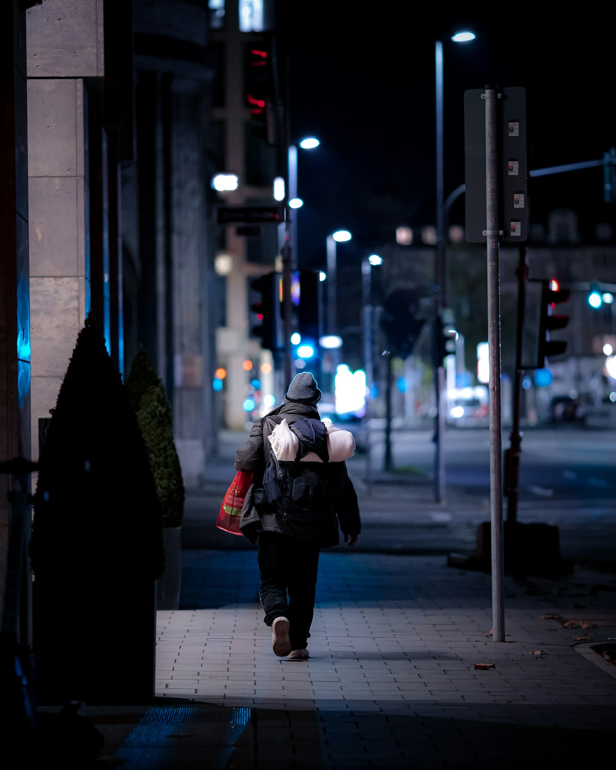 A person walking on a city sidewalk at night, carrying a bag and wearing a beanie and coat, with street lights illuminating the scene.