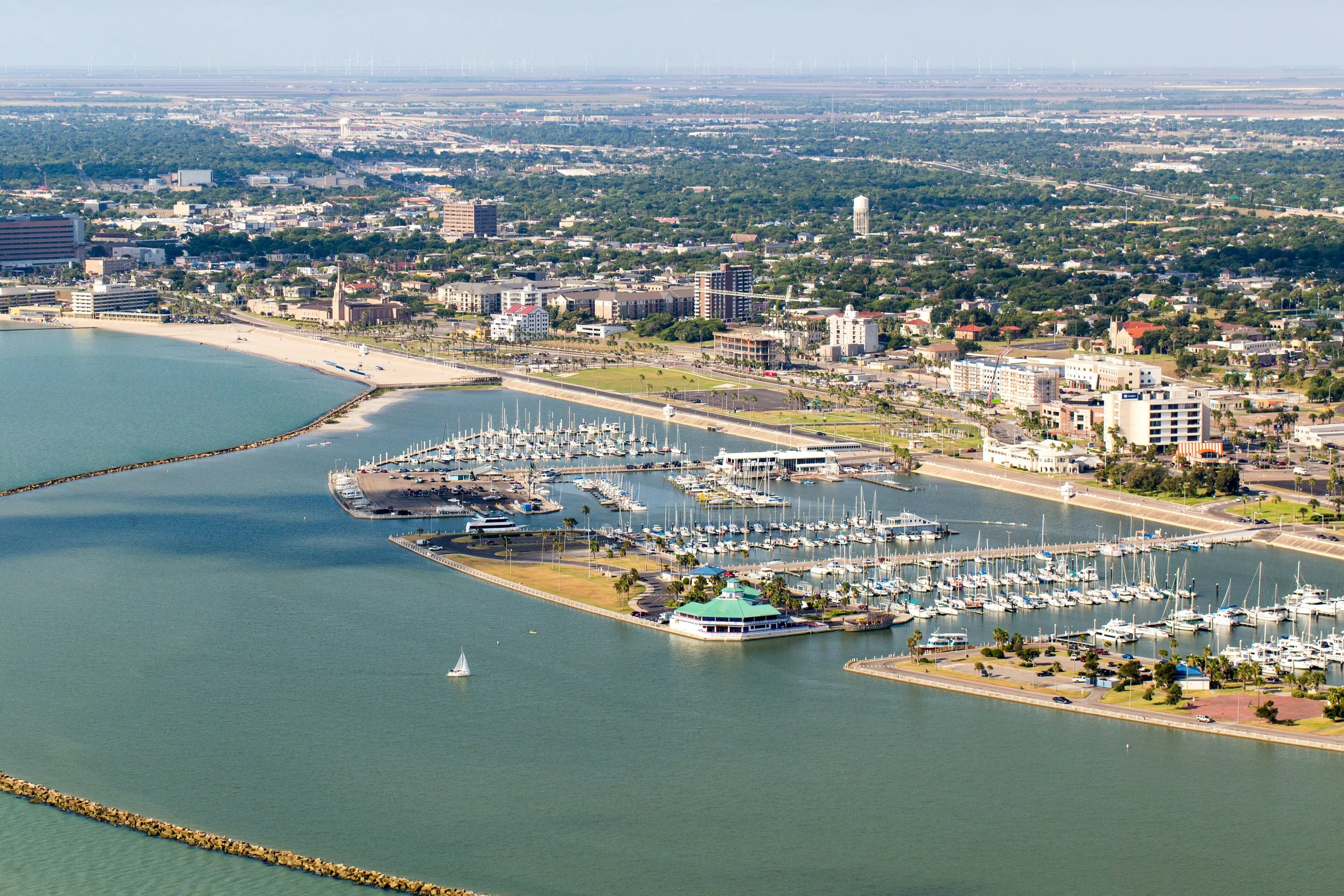 Aerial view of a marina with numerous sailboats docked, surrounding pathways, city buildings, and a beach shoreline with calm water.