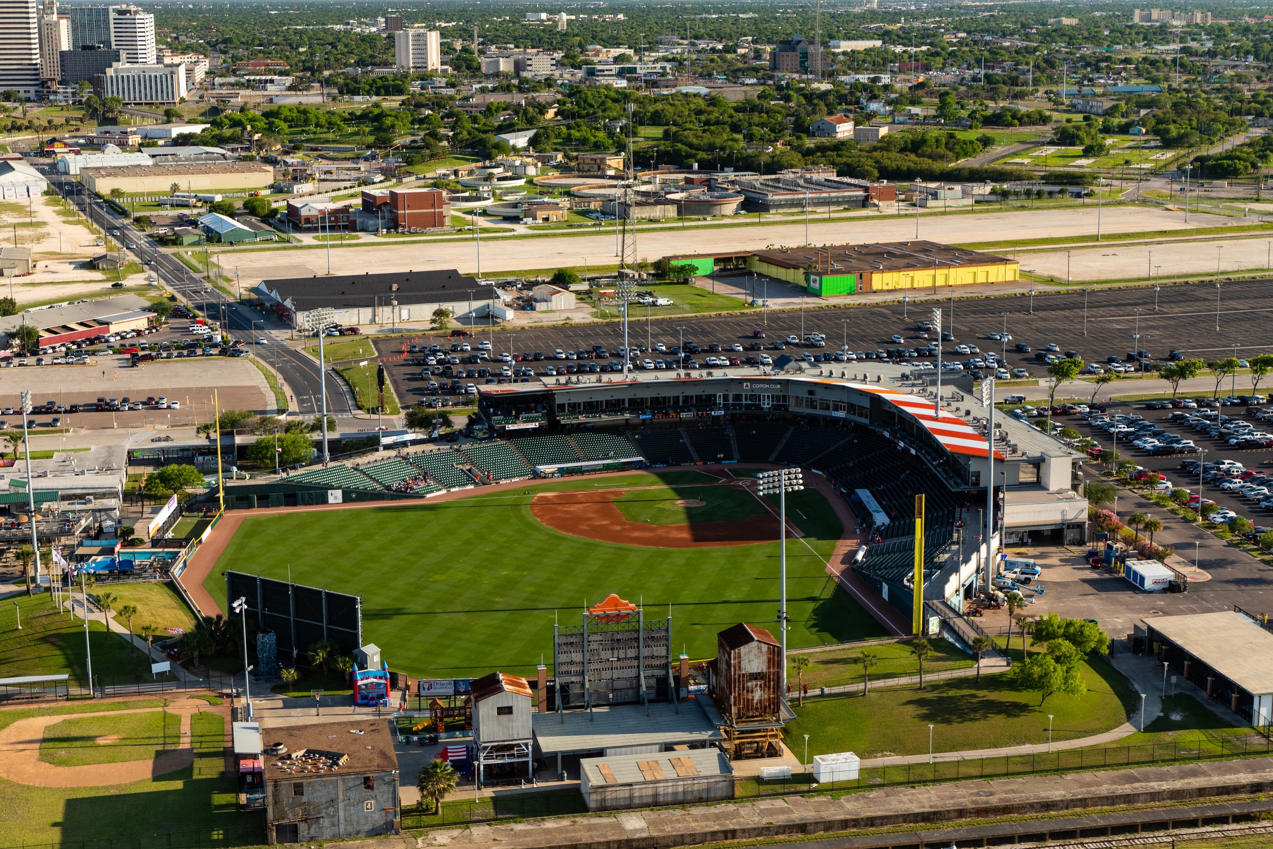 Aerial view of a sports stadium with a baseball field, surrounded by parking lots and city buildings in the background.