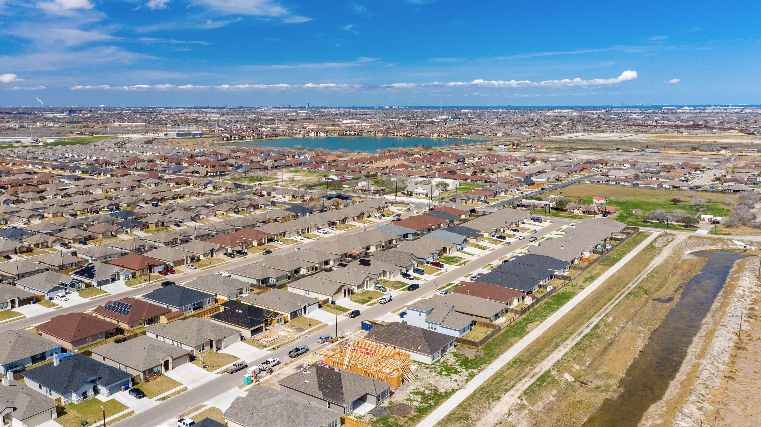 Aerial view of a residential neighborhood with single-family houses, a pond, and a clear blue sky with few clouds.