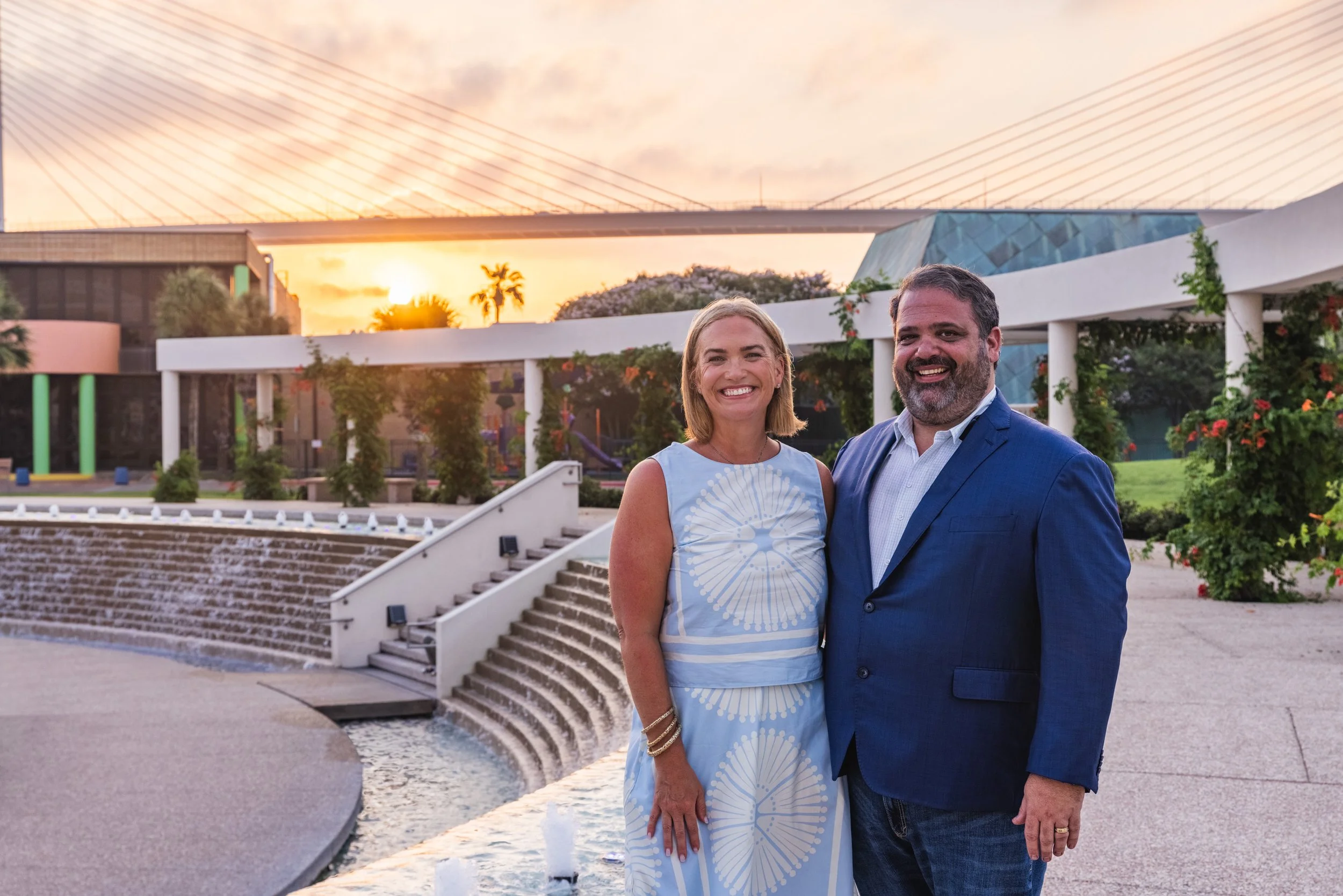 A smiling couple, a woman in a light blue dress with a white circular pattern and a man in a blue suit, standing outdoors near a water fountain with stairs, framed by greenery, during sunset.