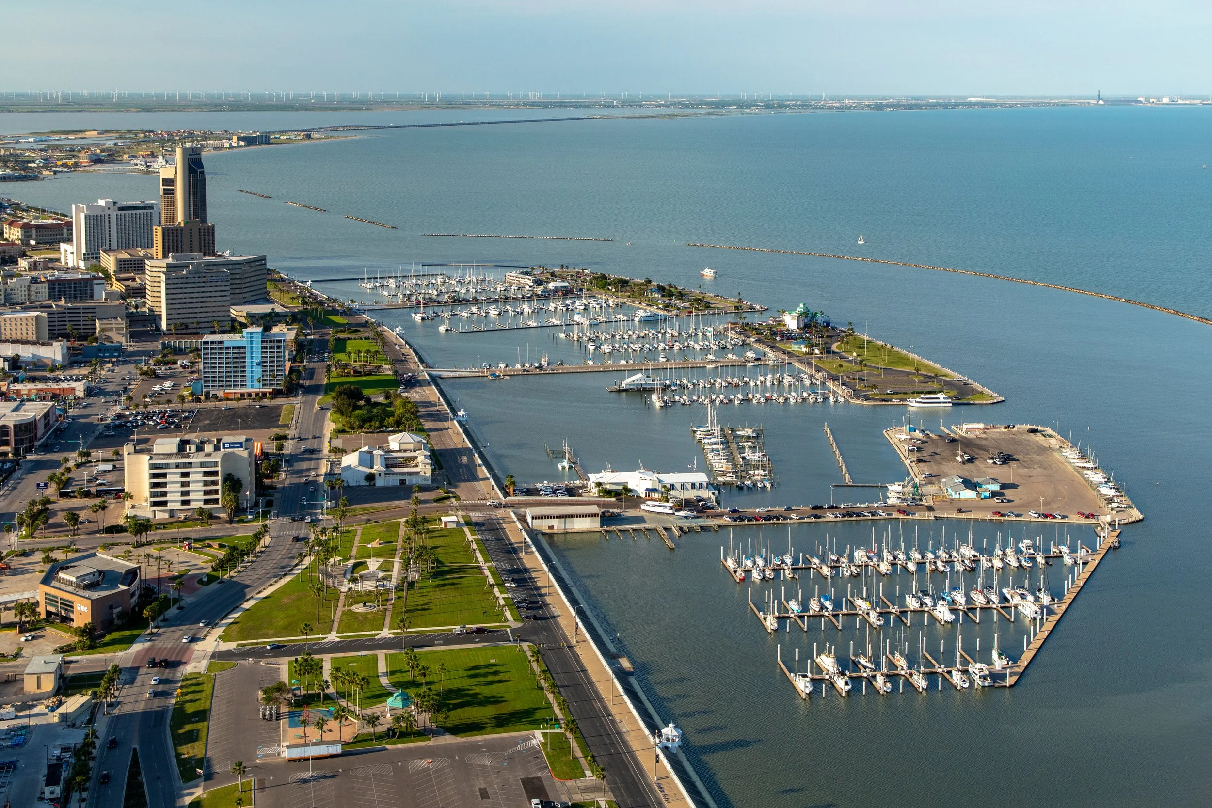 Aerial view of a coastal city with a marina, high-rise buildings, and a large body of water with sailboats and wind turbines in the distance.