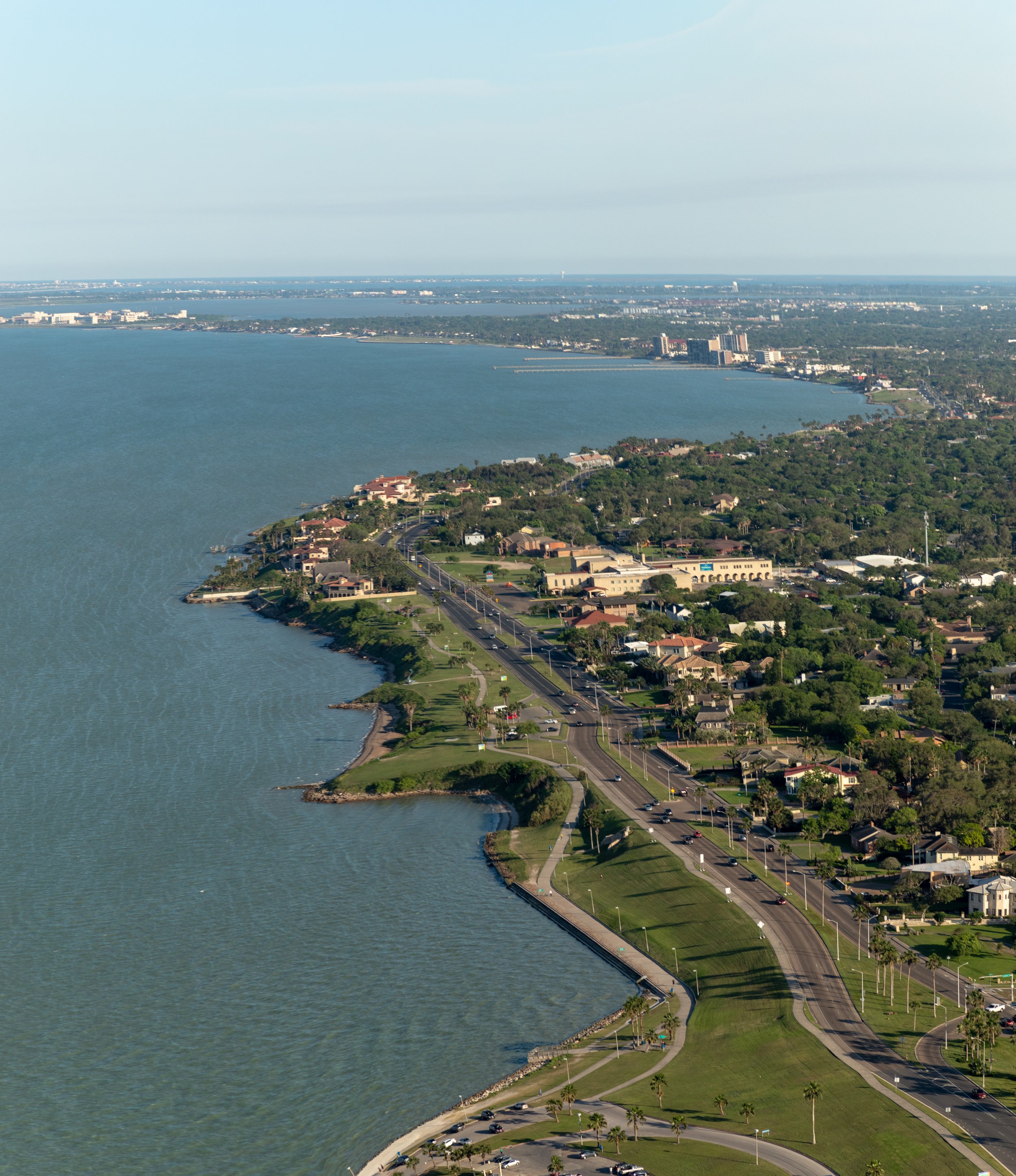 Aerial view of a coastal city with a large body of water on the left and green parkland with walking paths and palm trees on the right, featuring a mix of residential and commercial buildings and a highway running through the area.