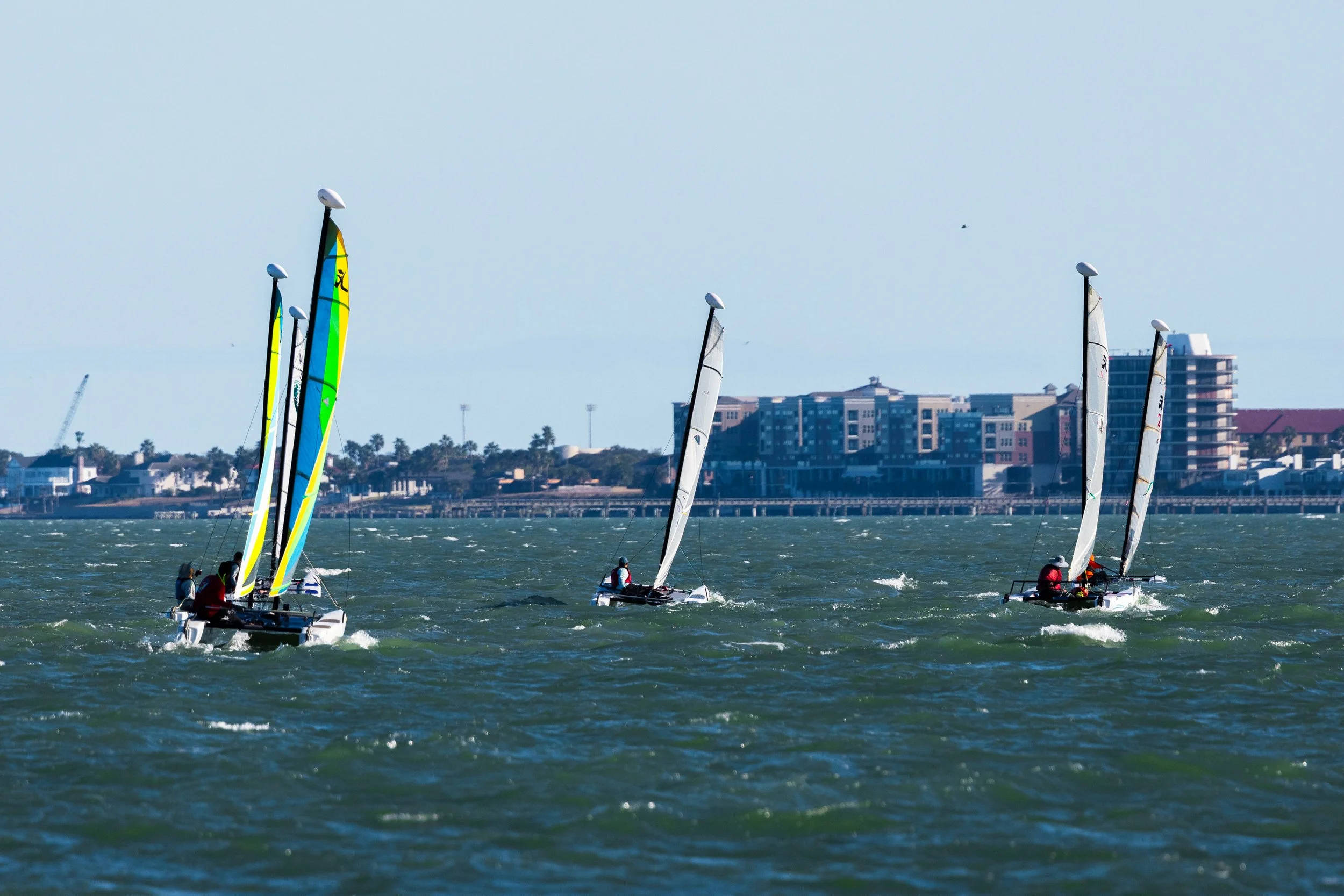 Multiple small sailboats with colorful sails on a body of water, with a cityscape in the background.