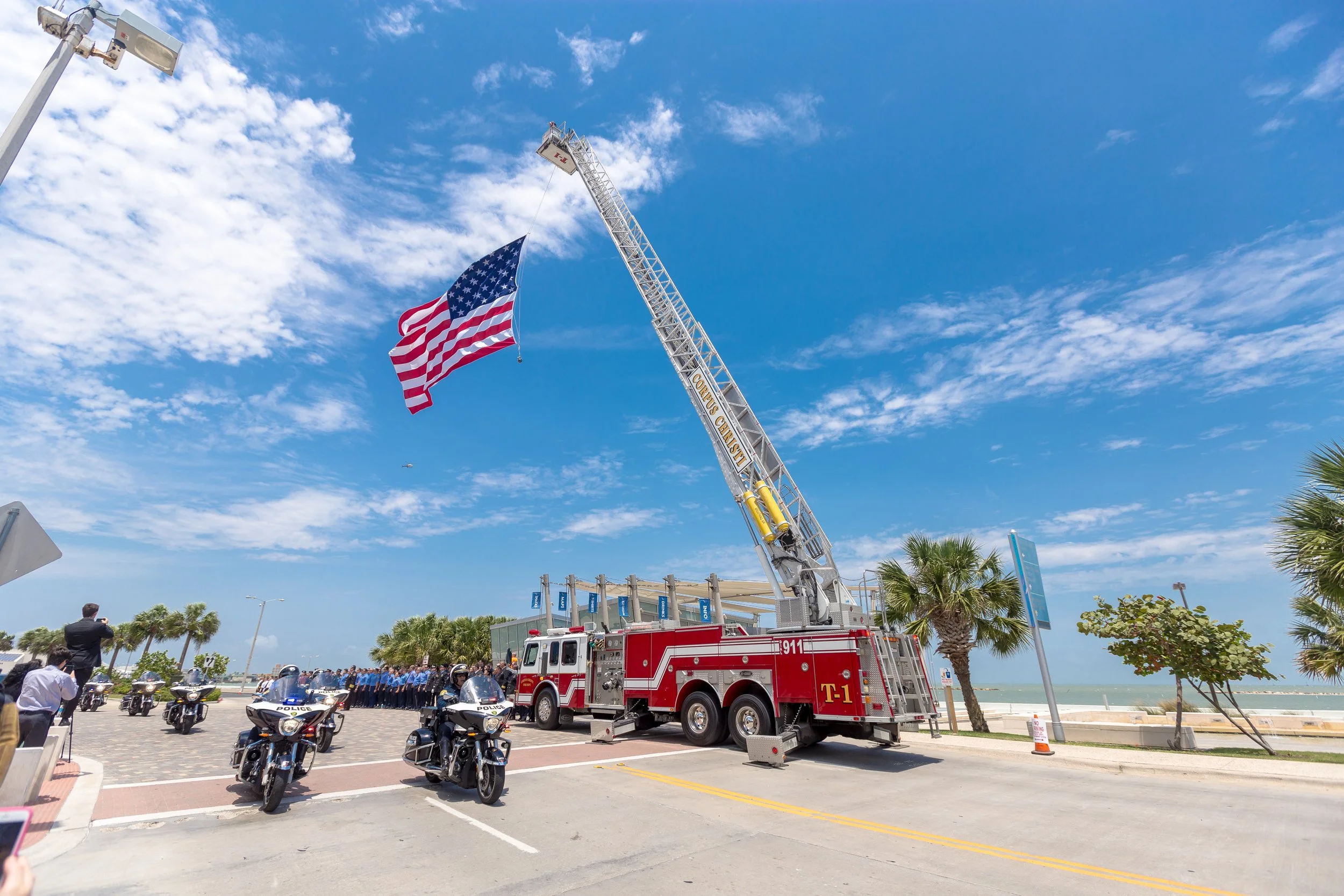 A fire truck with an extended ladder hoisting an American flag at a public event near the beach, lined with palm trees, with police motorcycles and officers in formation in the foreground, and a bright blue sky with scattered clouds overhead.